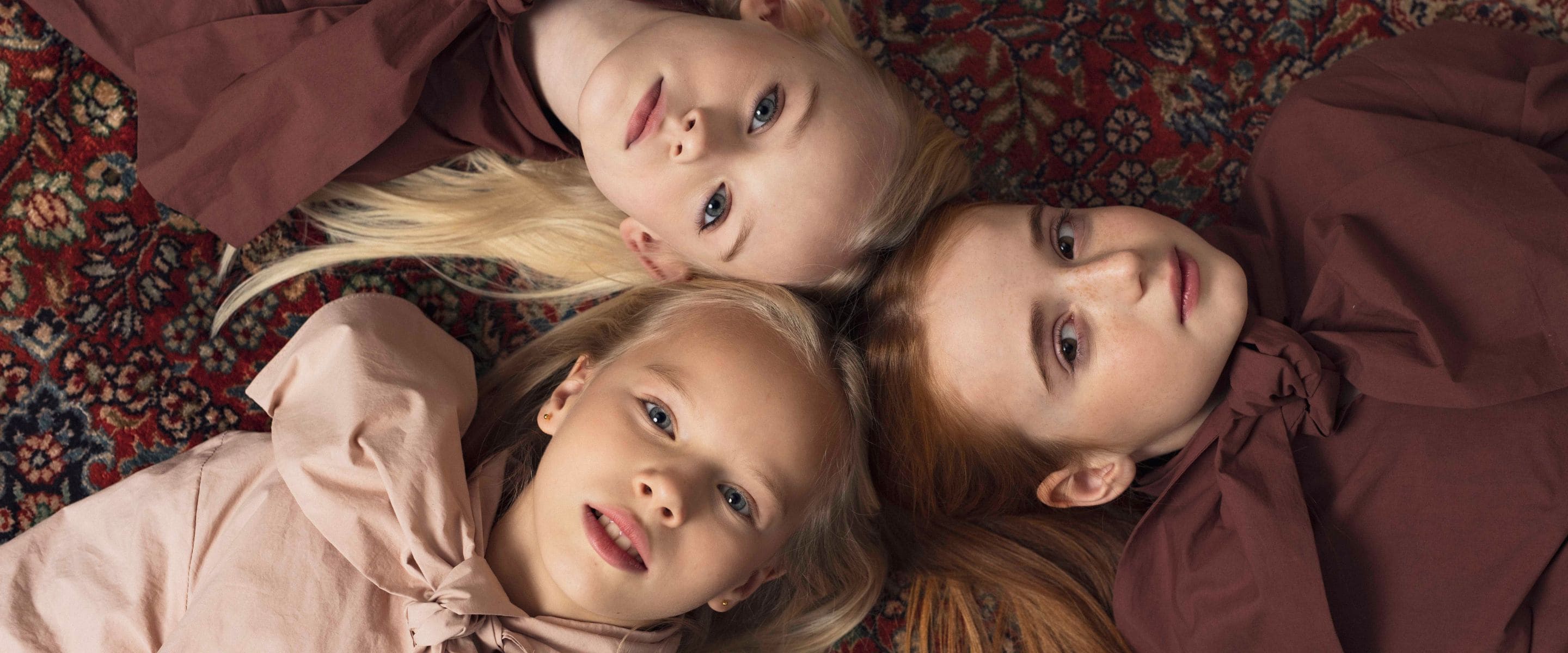 Three girls lying on a patterned rug, looking at the camera, wearing coordinated dusty pink and mauve outfits.
