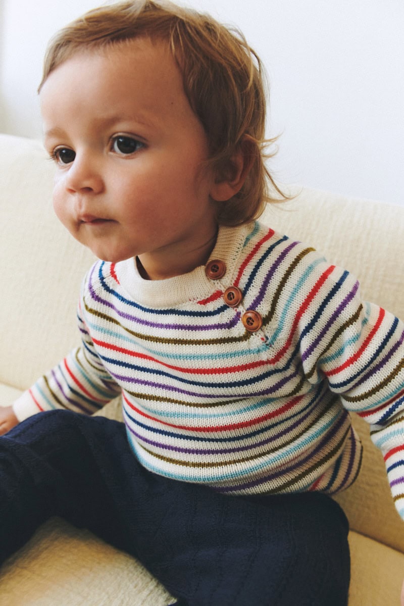 Toddler in a colorful striped knit sweater and dark trousers sitting on a cream sofa.