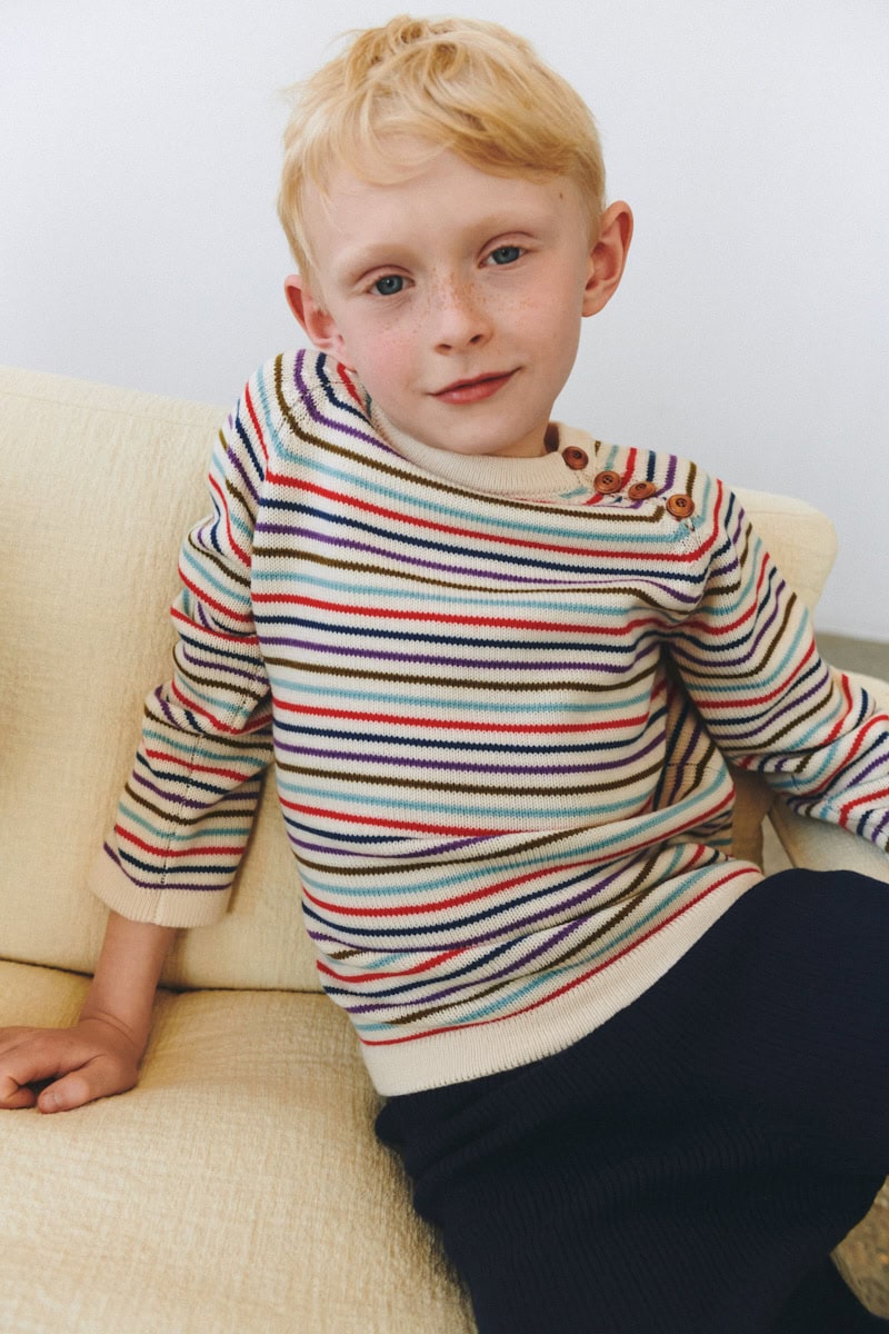 Young boy wearing a multicoloured striped FUB knit sweater with wooden buttons, sitting on a cream sofa.
