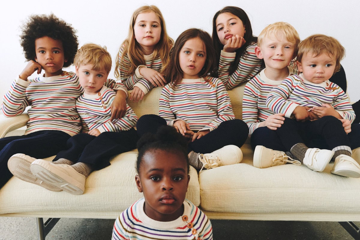 Group of children wearing matching multicoloured striped FUB knit sweaters, sitting together on a cream sofa in a bright interior.