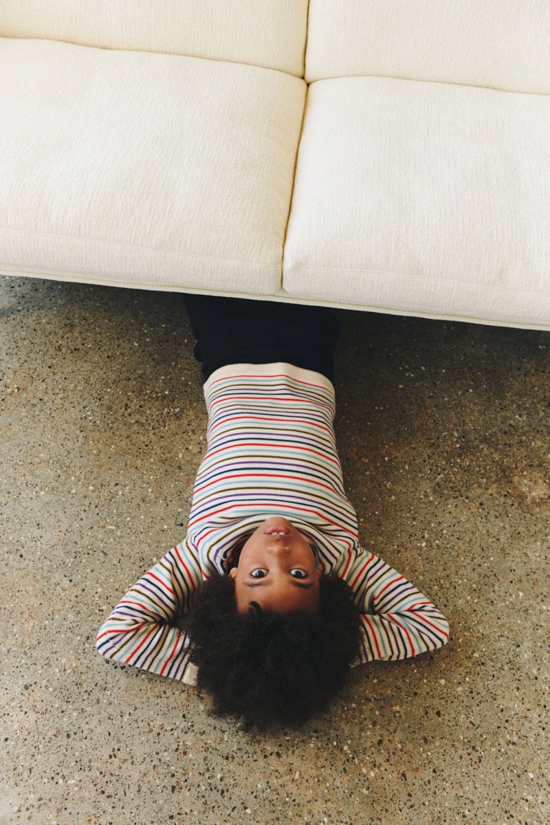 Child lying upside down on the floor beneath a cream sofa, wearing a cream knitted sweater with multicolored horizontal stripes and dark trousers.