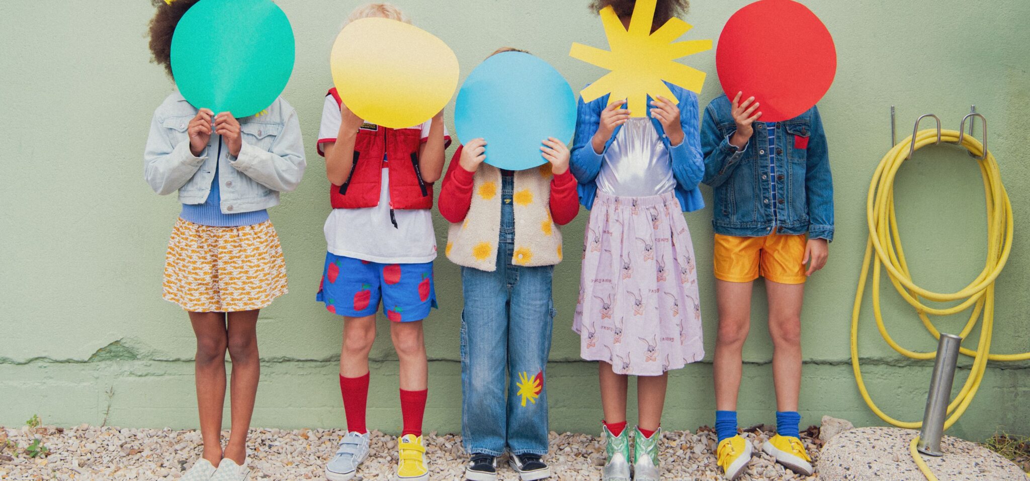 Kids fashion editorial RE.Twist photographed by Emmanuelle Lhote, featuring children wearing colourful upcycled clothes and holding graphic shapes in front of a green wall for Scimparello Magazine.