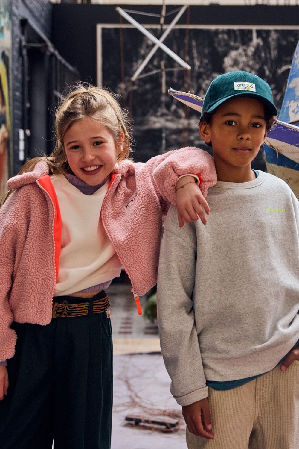 Two children posing beside a vintage boat sculpture wearing cozy fleece jackets, sweatshirt and casual AW26 clothing.