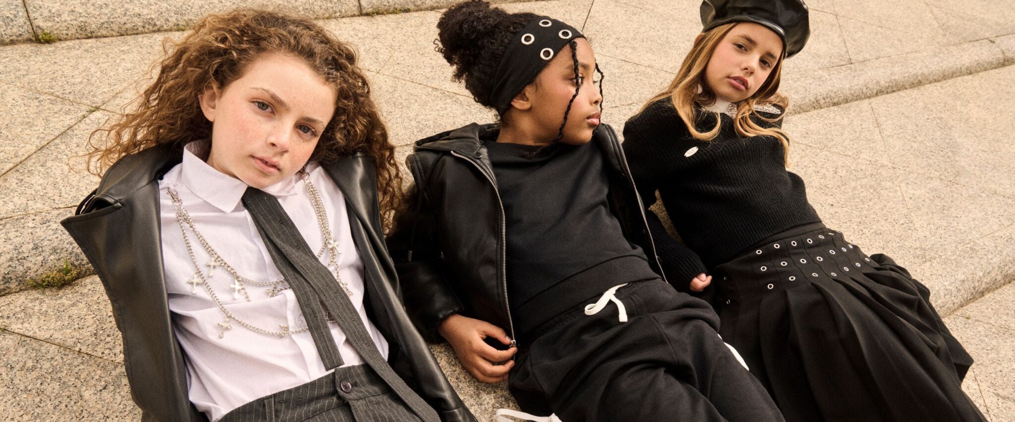 Three girls in coordinated black kidswear looks pose together on urban steps, wearing a leather jacket, pleated skirt, knitwear, statement accessories and a black beret.