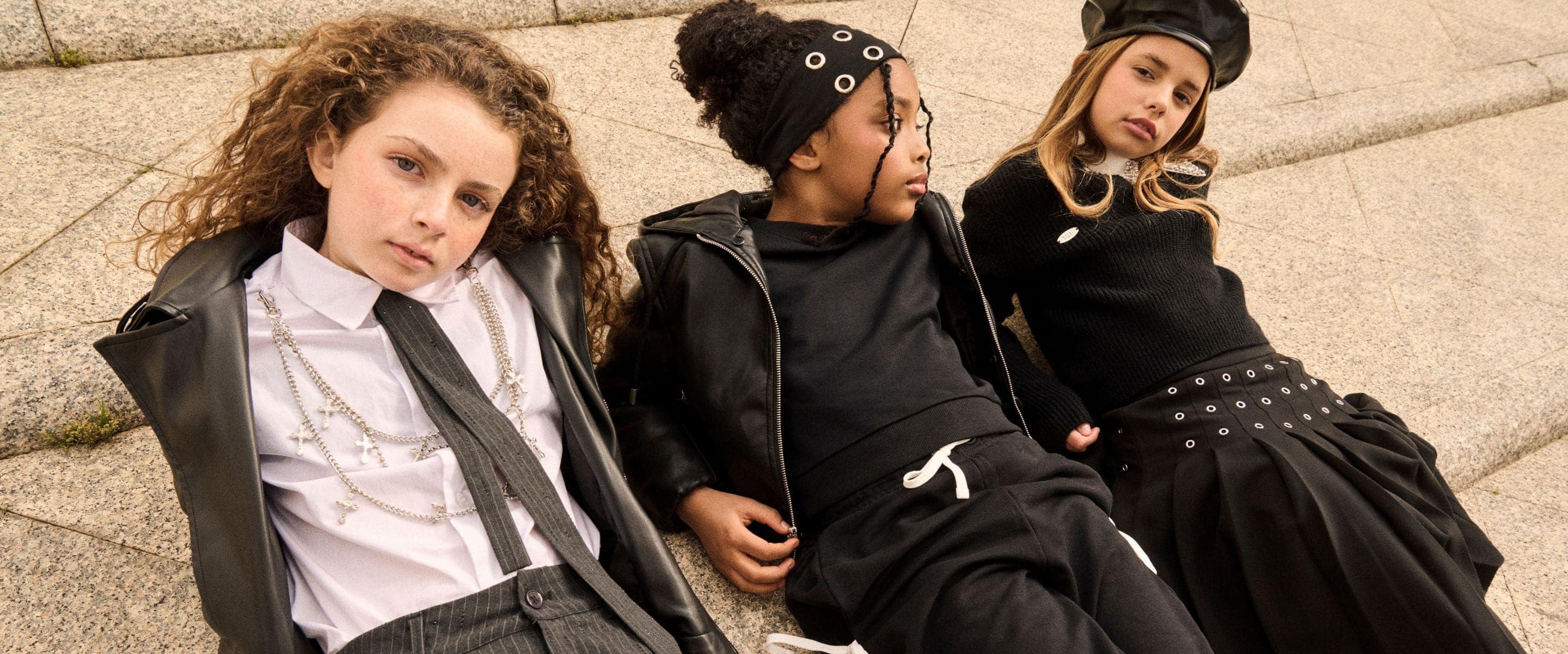 Three girls in coordinated black kidswear looks pose together on urban steps, wearing a leather jacket, pleated skirt, knitwear, statement accessories and a black beret.