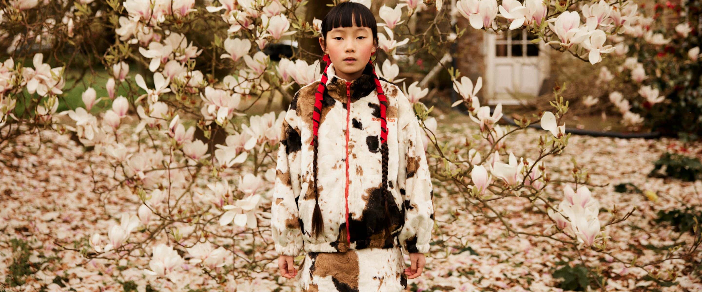 A young girl wearing a cow-print fleece jacket and matching skirt stands among blooming magnolia trees in a garden.