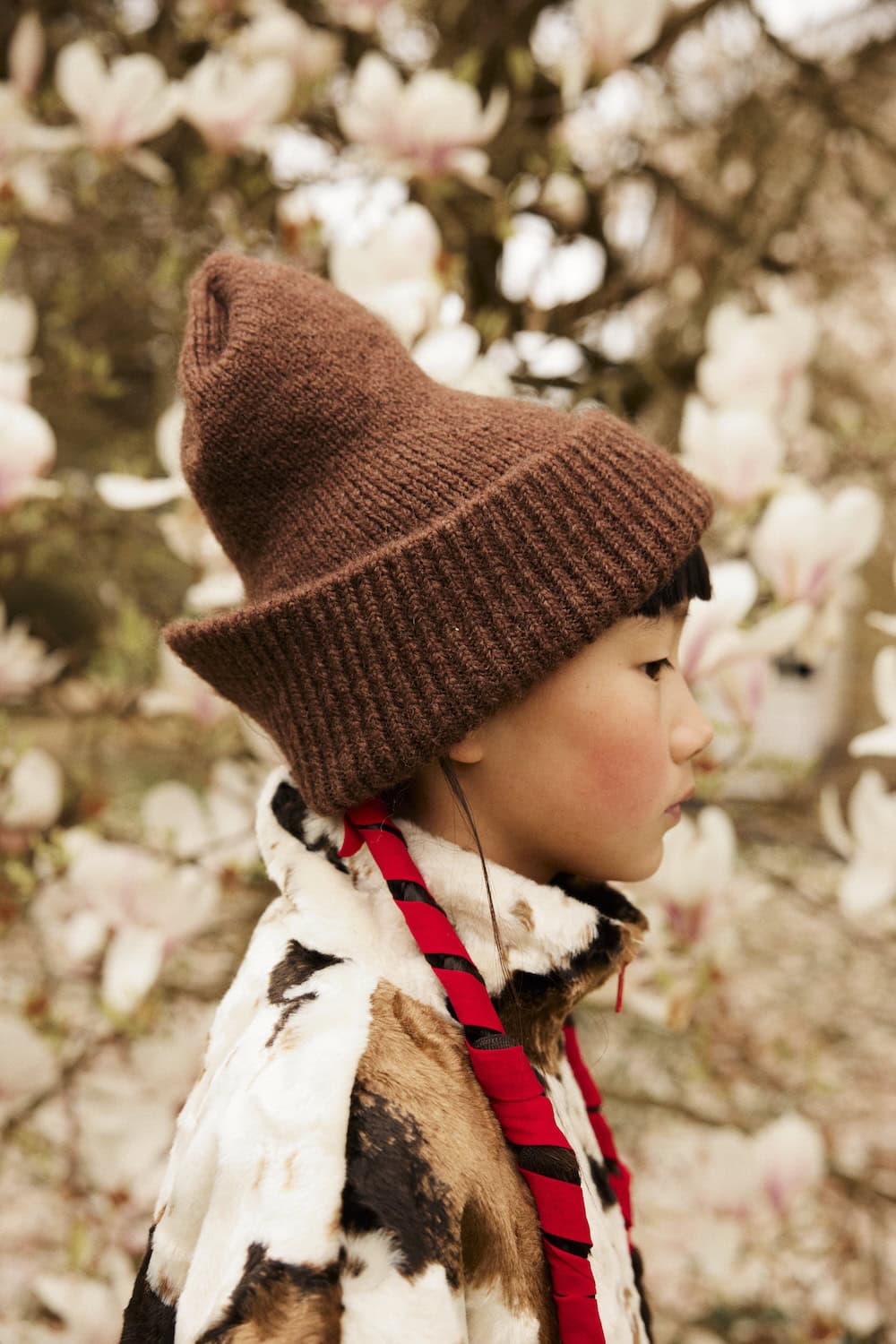 Close-up of a young girl wearing a brown knit cat-ear beanie and cow-print fleece jacket among magnolia blossoms.