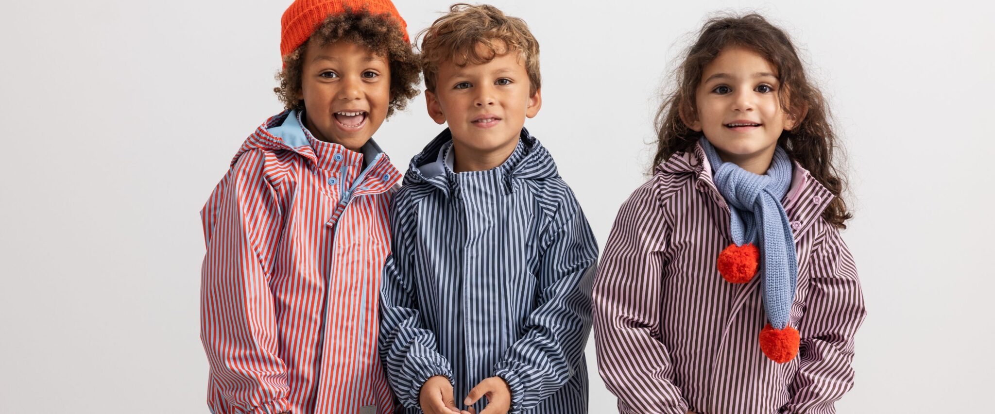 Three smiling children wearing colorful striped waterproof outfits, standing together against a white background.