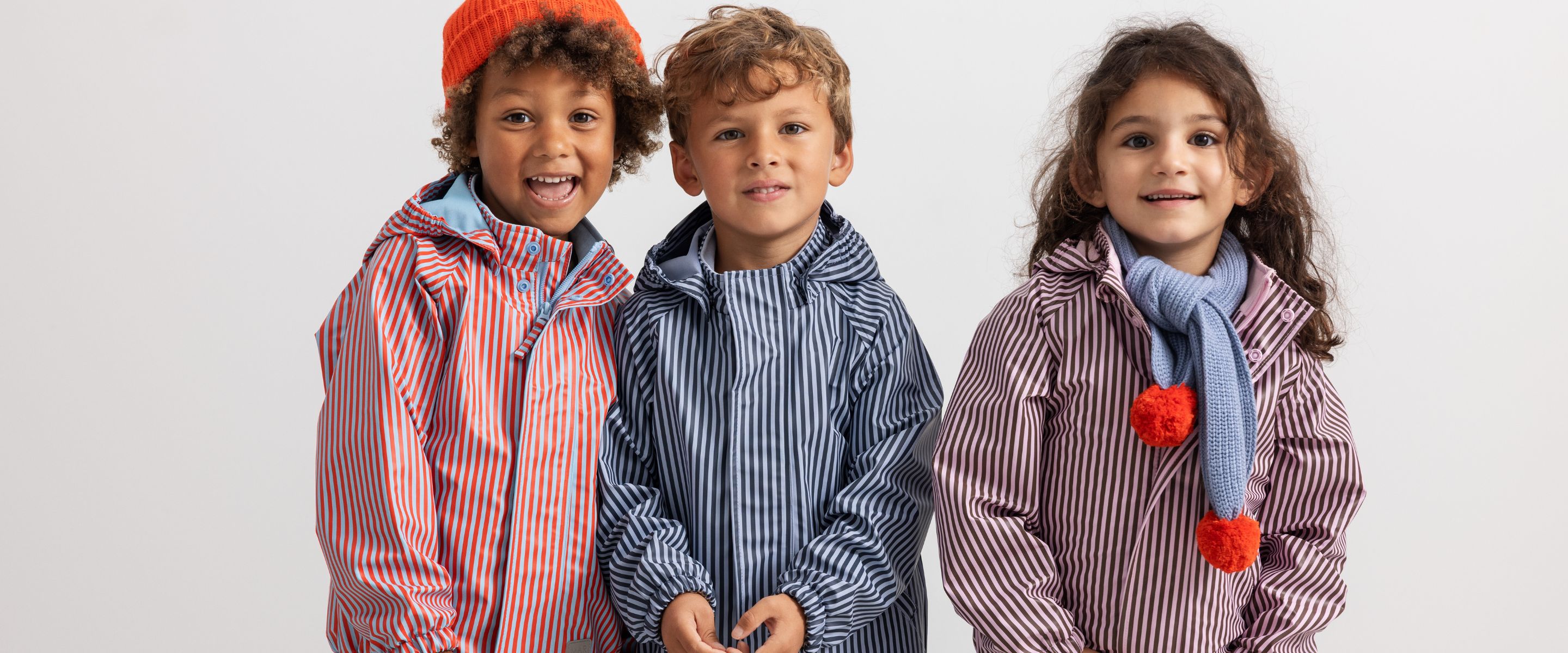 Three smiling children wearing colorful striped waterproof outfits, standing together against a white background.