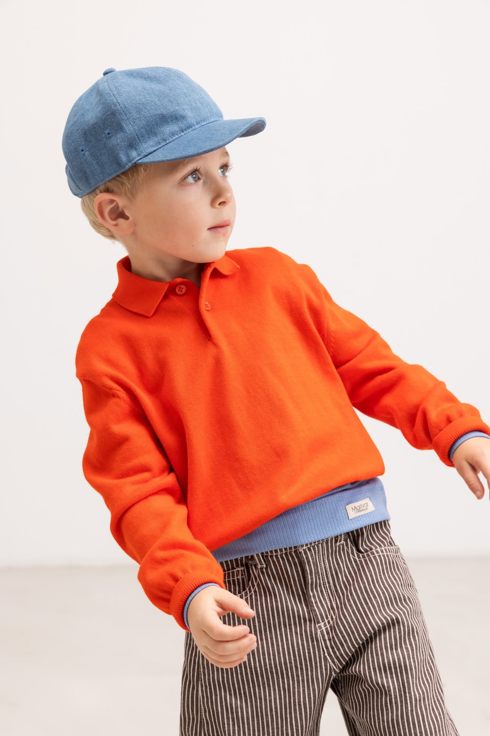 Boy wearing an orange long-sleeve polo shirt, blue cap and brown striped trousers from MarMar Copenhagen FW26.