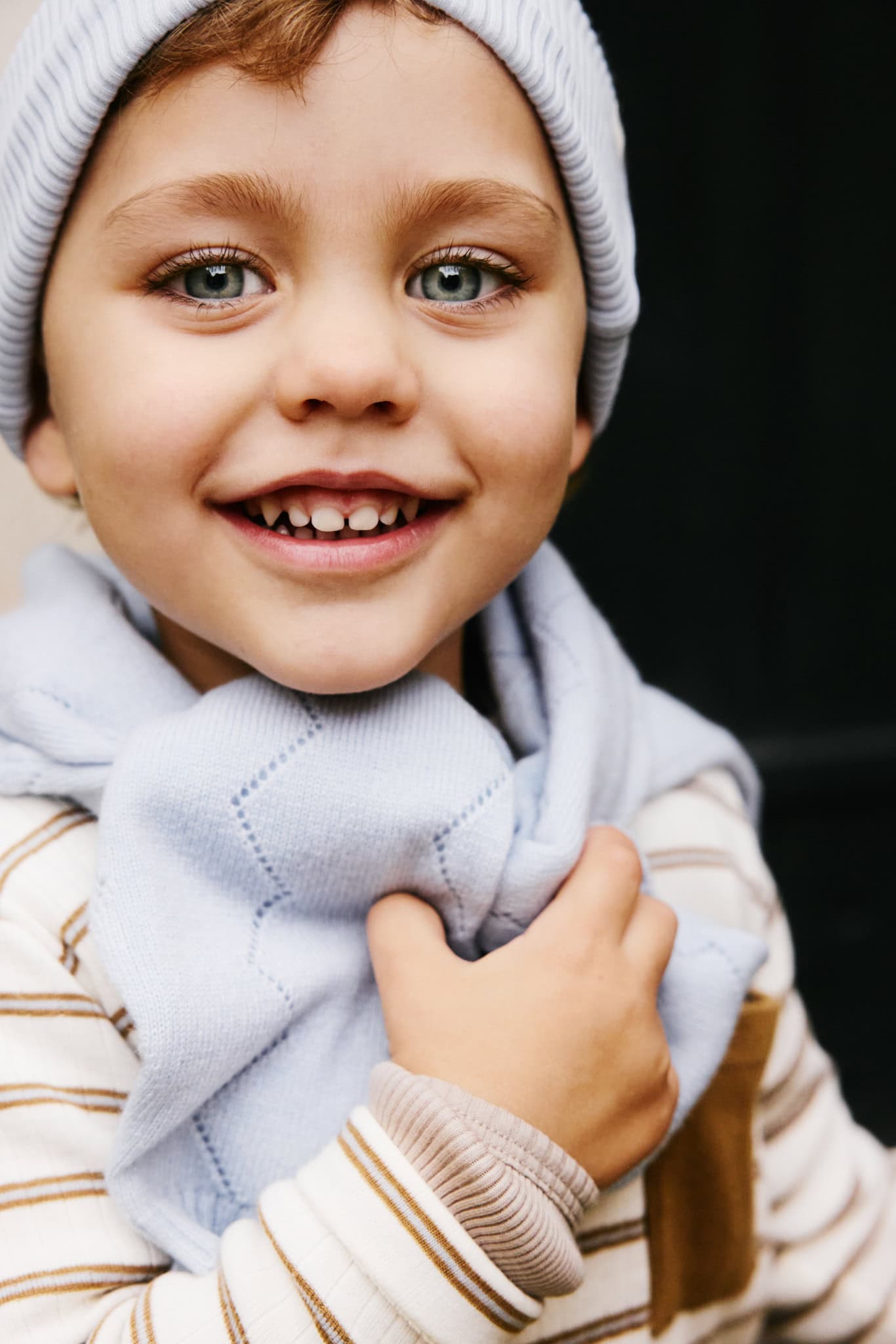 Smiling child wearing a light blue knitted hat, scarf and striped sweater from the Minimalisma FW26 collection.