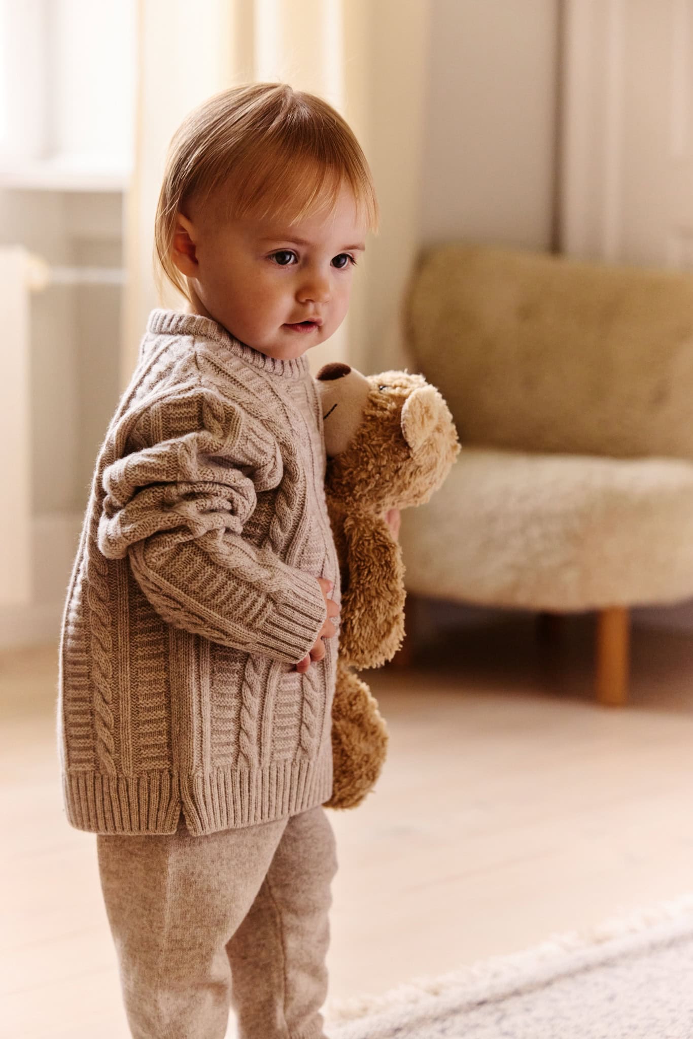 Toddler wearing a chunky cable-knit cardigan and trousers, holding a teddy bear in a cosy interior for the Minimalisma FW26 campaign.