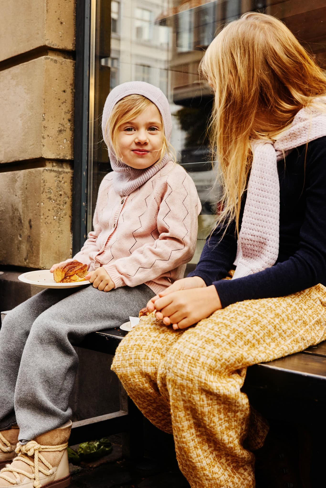 Child wearing a pink knitted cardigan and beanie, sitting with another child in Copenhagen as part of the Minimalisma FW26 campaign.