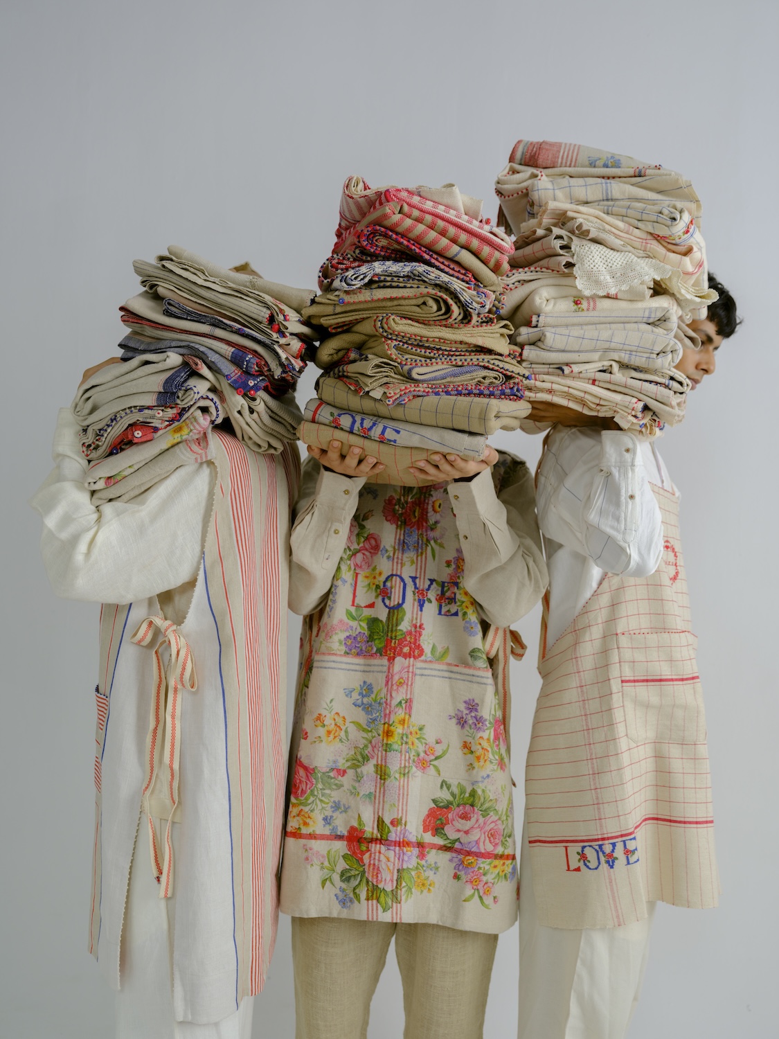 Three people carrying stacks of handwoven and embroidered fabrics from the péro Home collection, featuring floral prints and delicate checks.