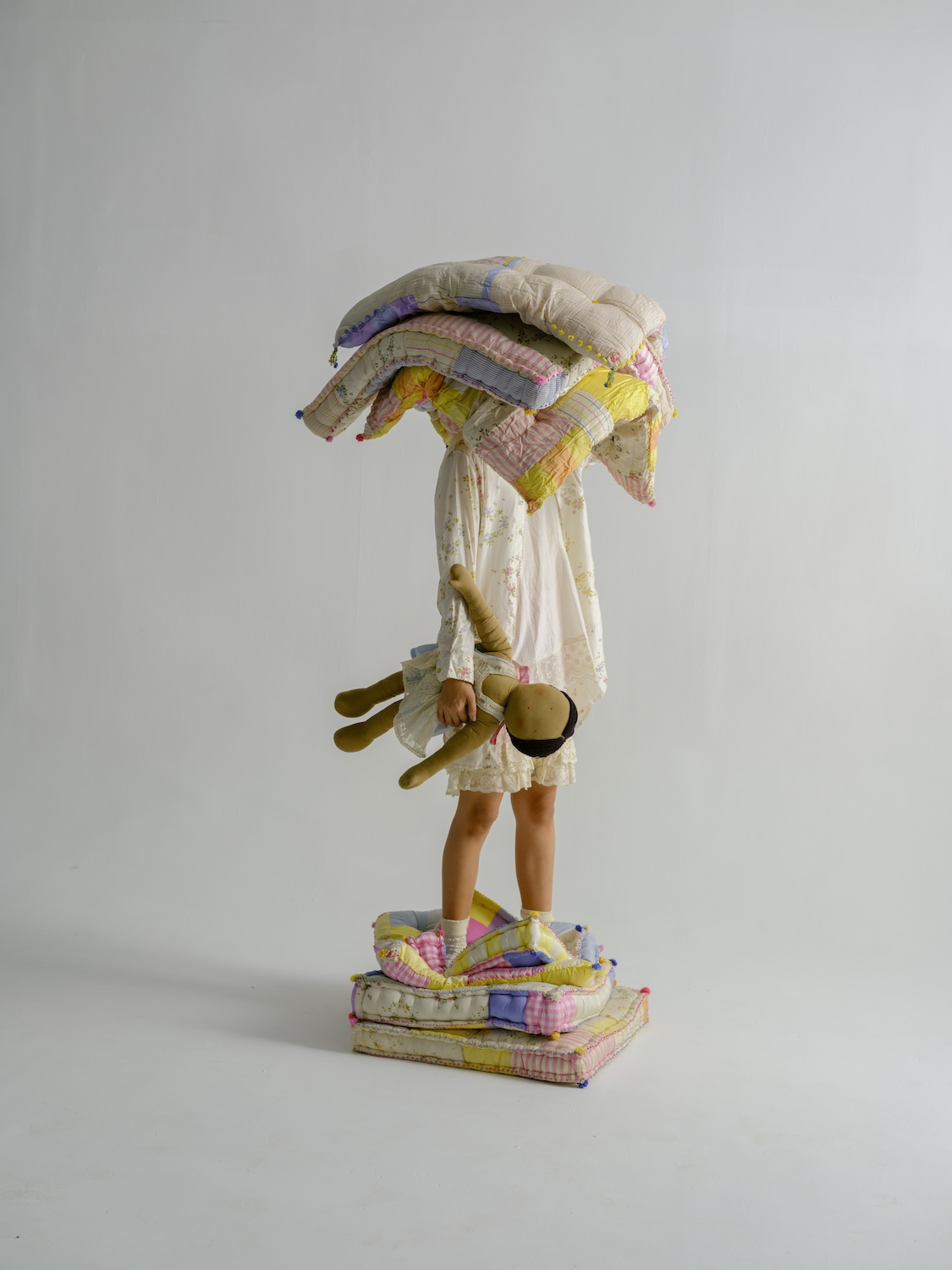Child holding a handmade rag doll and balancing a stack of colourful patchwork quilts on their head, wearing a floral outfit from péro Home.