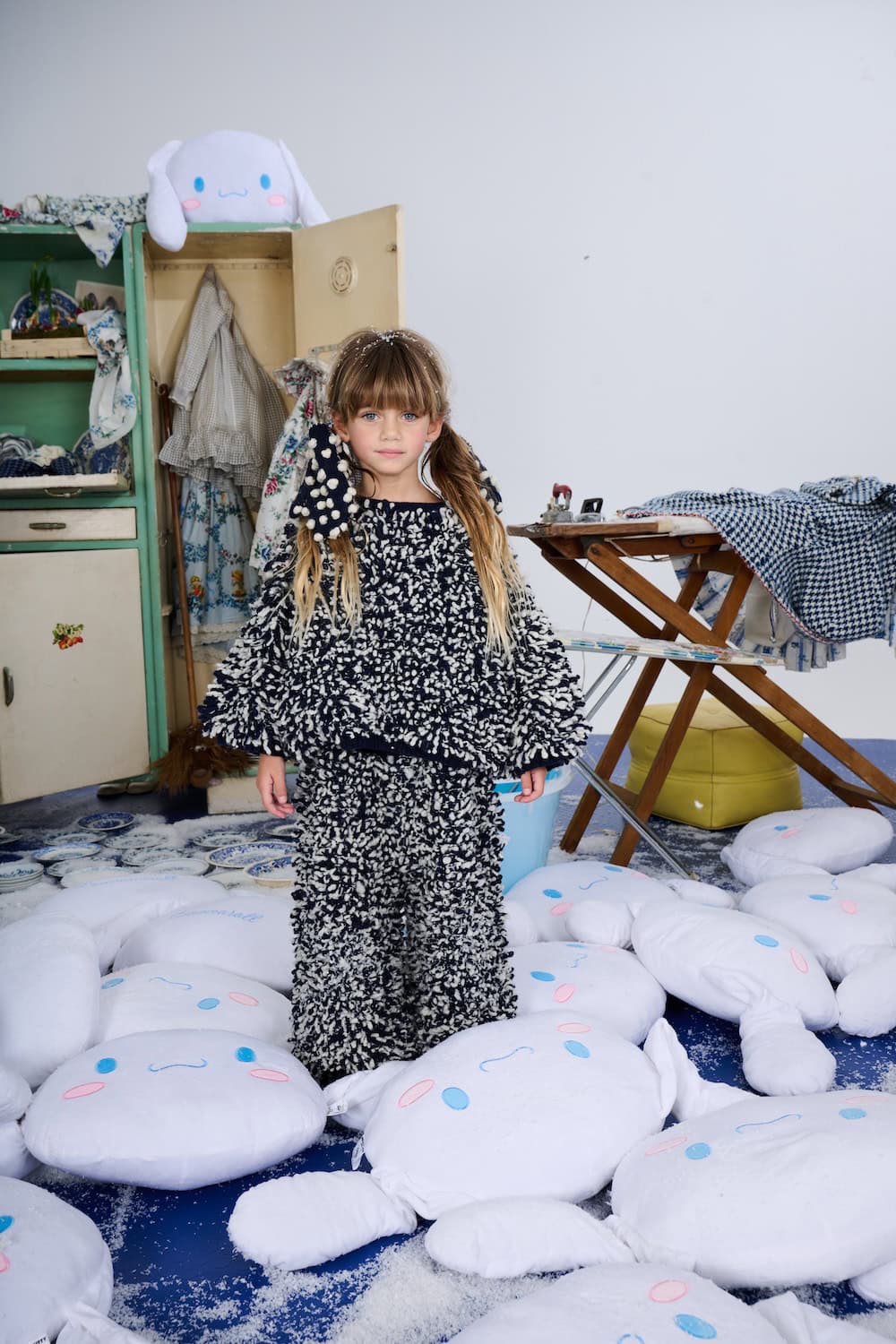 Girl wearing a navy and white textured knit sweater and trousers standing among oversized white cushions in a snowy room.