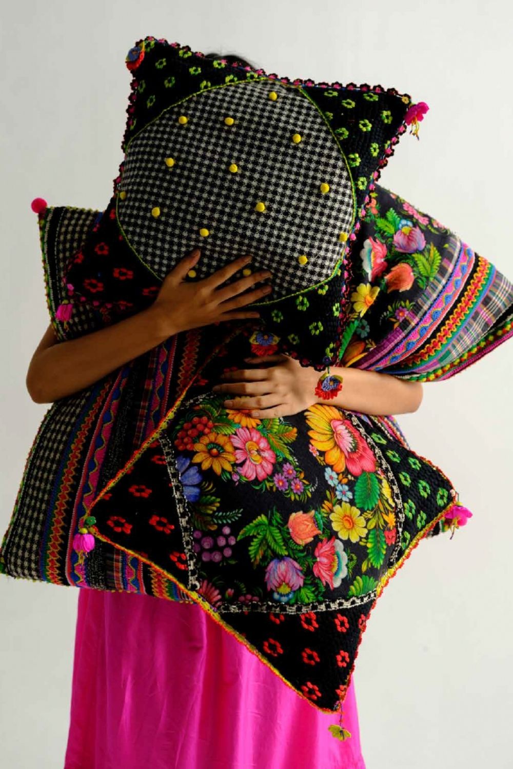 Person surrounded by colourful embroidered and floral patchwork cushions from the péro Home collection, wearing a bright pink outfit.