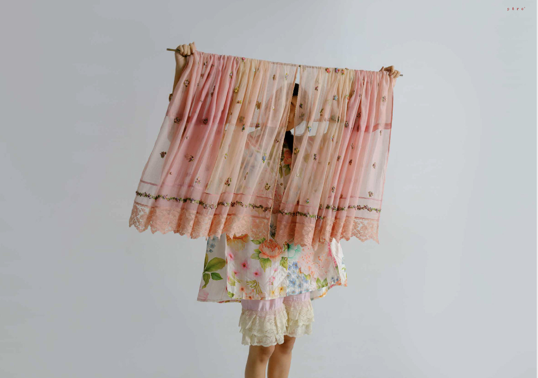Person holding a sheer pink embroidered curtain from the péro Home collection, revealing colourful floral textiles underneath.