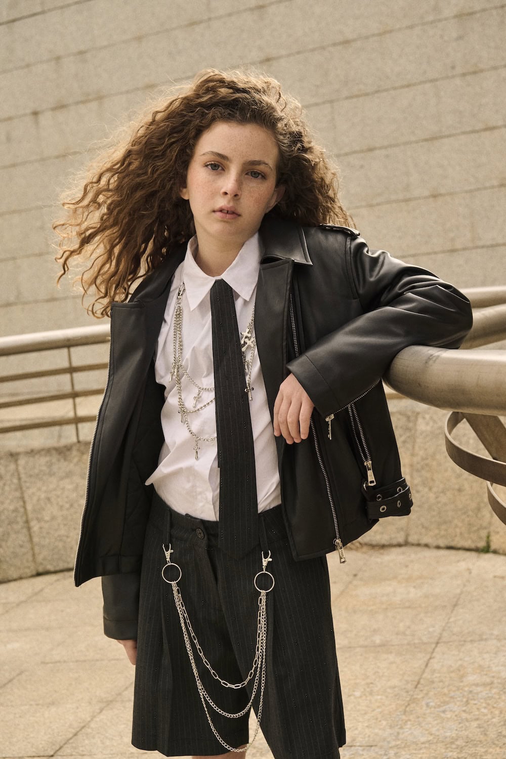 Young girl wearing a black leather jacket, white shirt and pinstripe trousers with chain details, posing outdoors in a modern urban setting.