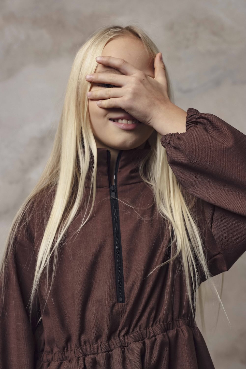 A blonde girl smiling and covering her eyes with one hand, wearing a brown zip-up jacket with gathered details, against a neutral textured background.