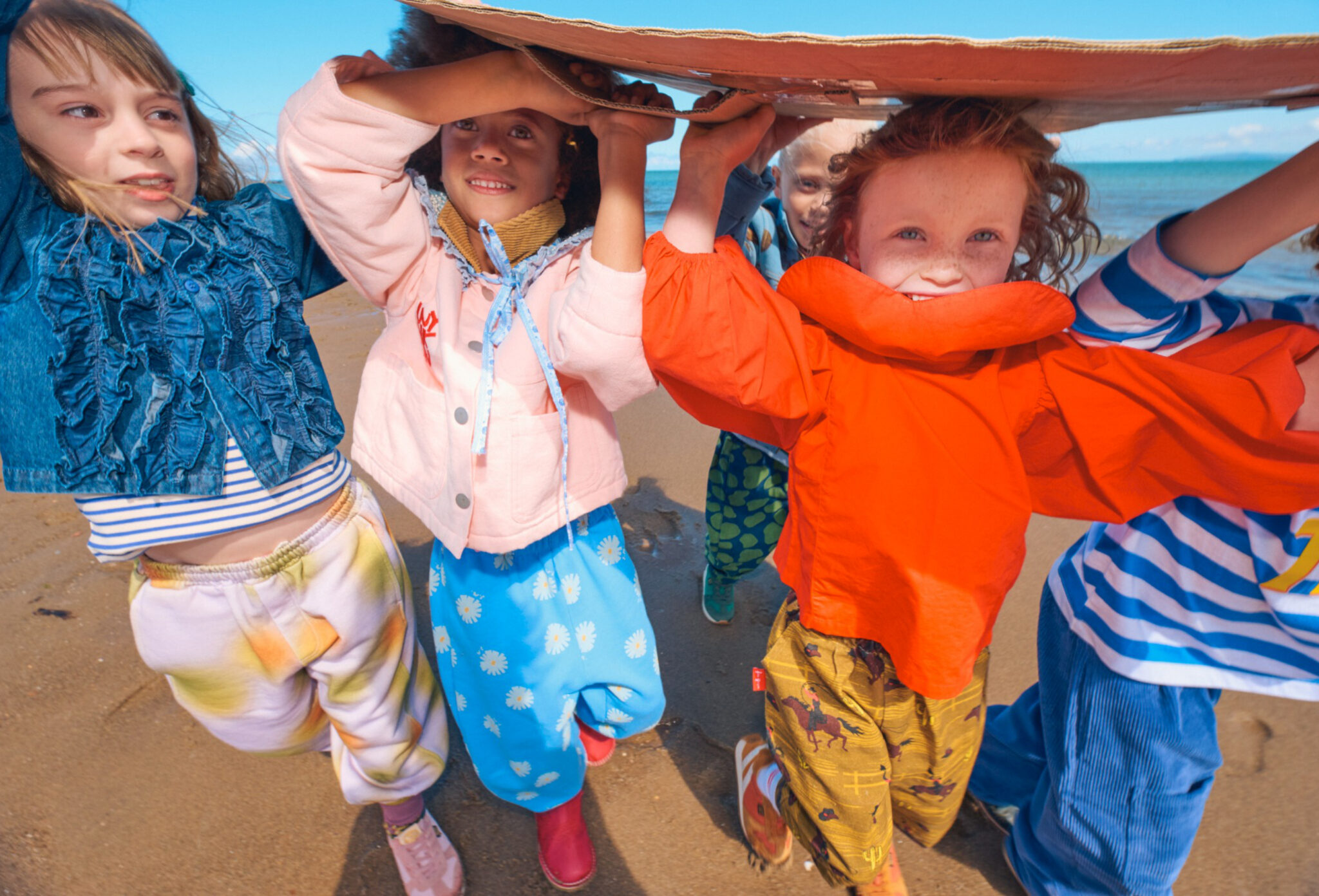 Forecast: Colour FW26 fashion editorial photographed by Alma Rosaz, featuring children in vivid colourful kids fashion looks running together on the sand under a bright blue sky for Scimparello Magazine.