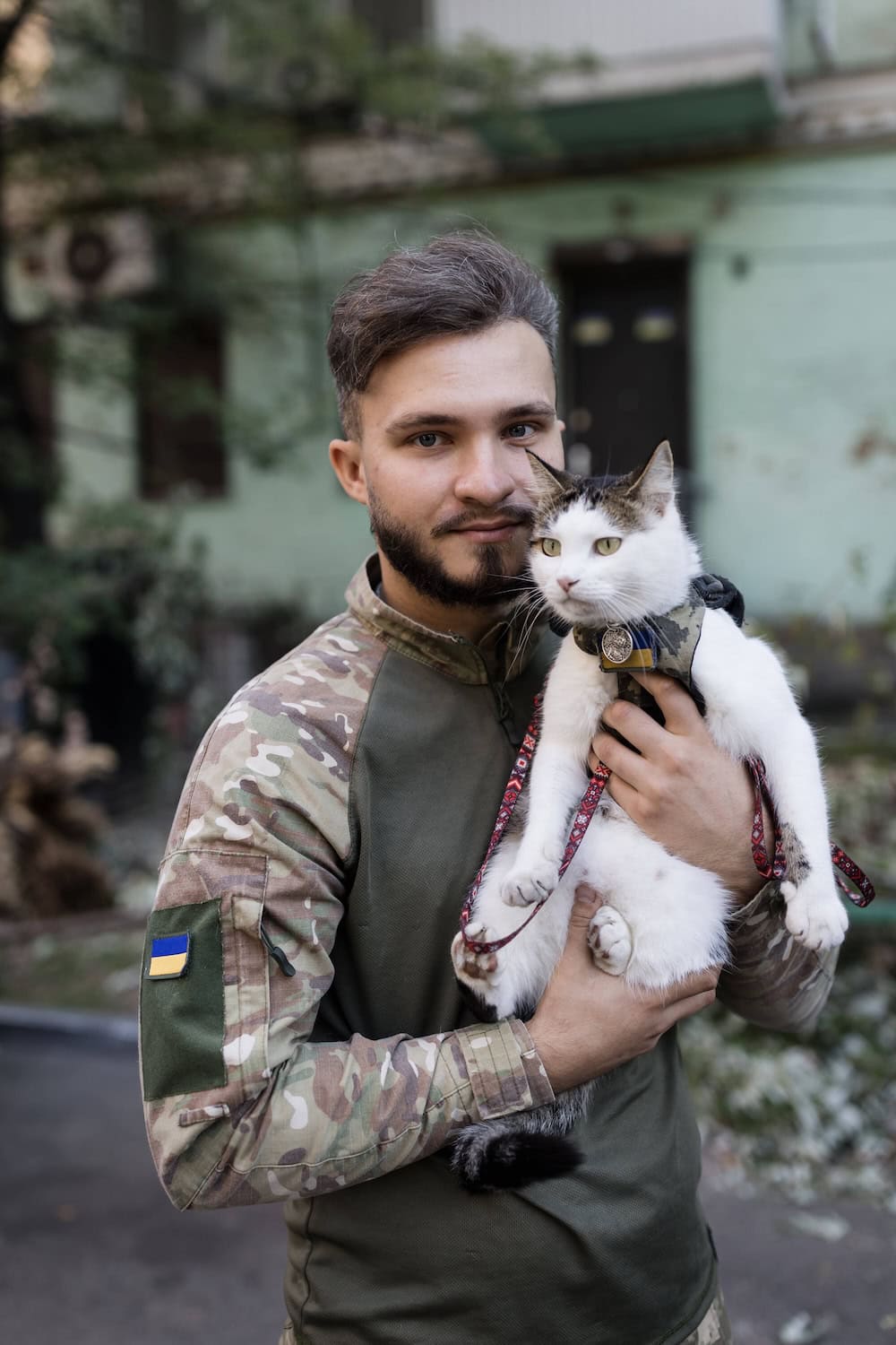 A Ukrainian soldier holding a white and grey cat wearing a harness, photographed outdoors in Ukraine.