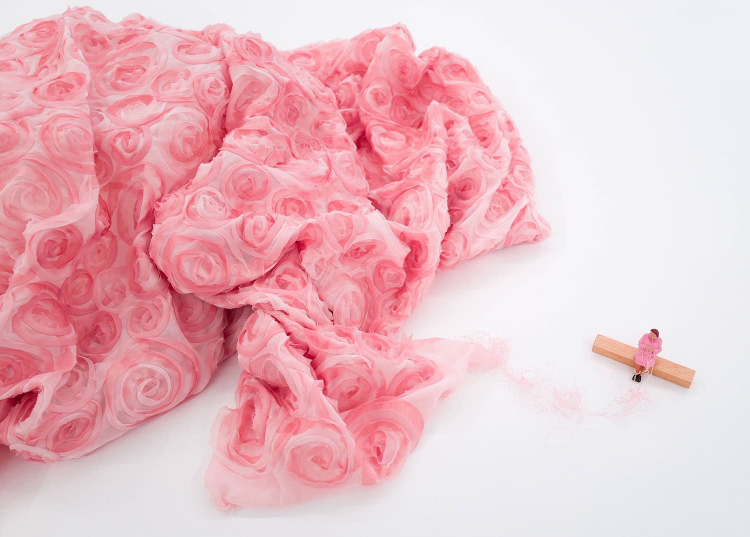 Liliana Porter’s “The Weaver” (2018), featuring a tiny figure sitting on a wooden beam beside a large mound of delicate pink rose-shaped fabric.