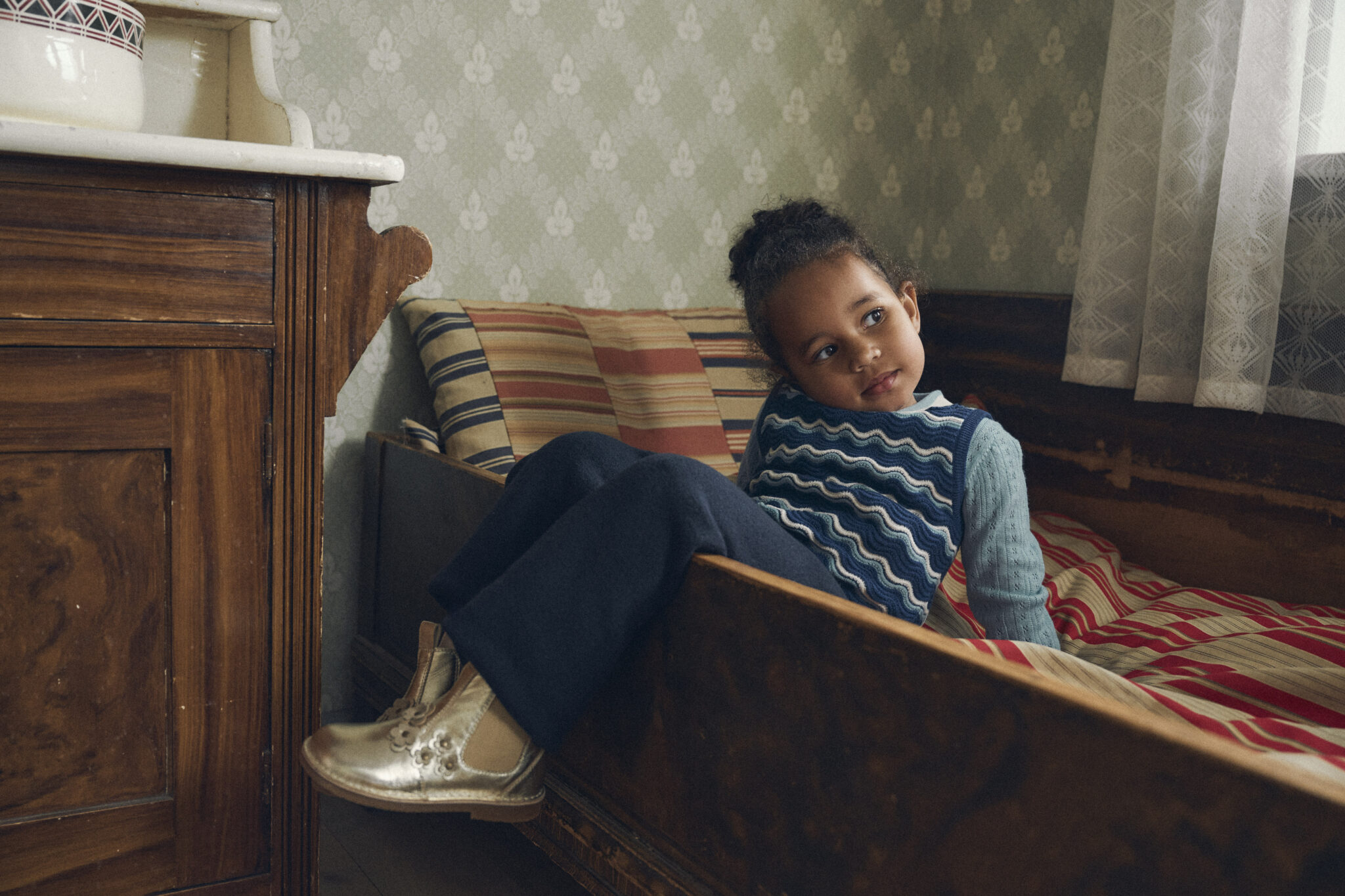 A child wearing a blue knit sweater and dark trousers sits on a vintage wooden bed, wearing silver Mary Jane shoes.