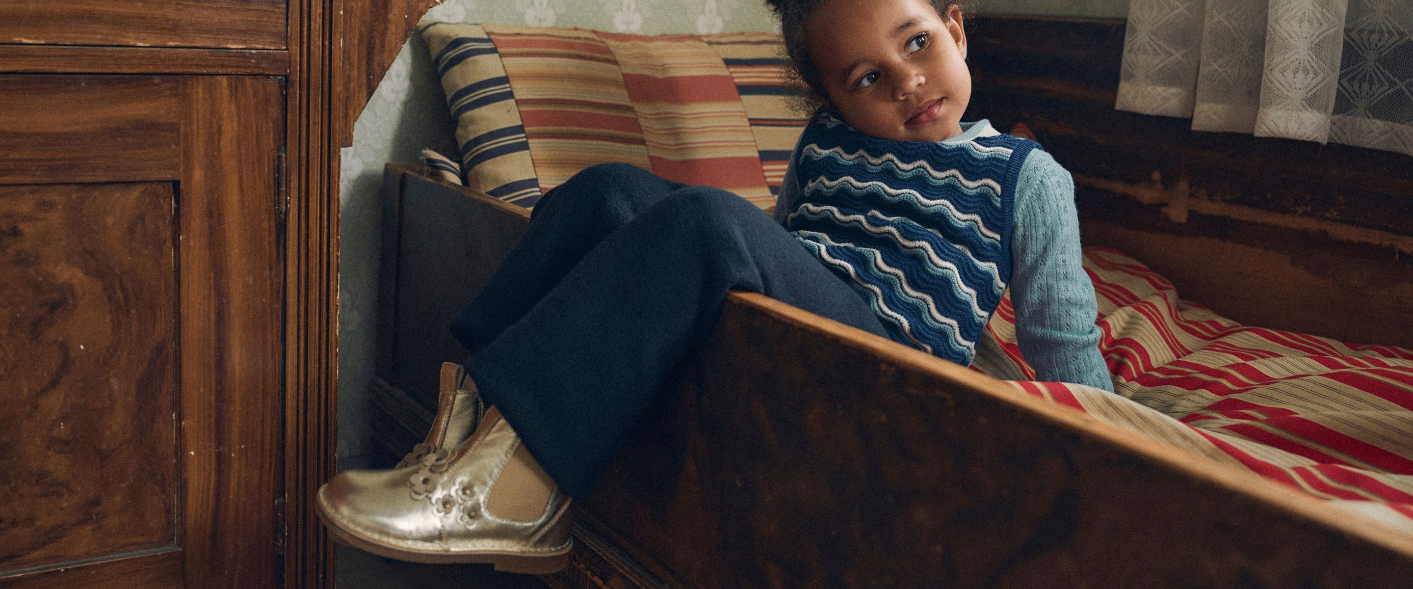 A child wearing a blue knit sweater and dark trousers sits on a vintage wooden bed, wearing silver Mary Jane shoes.