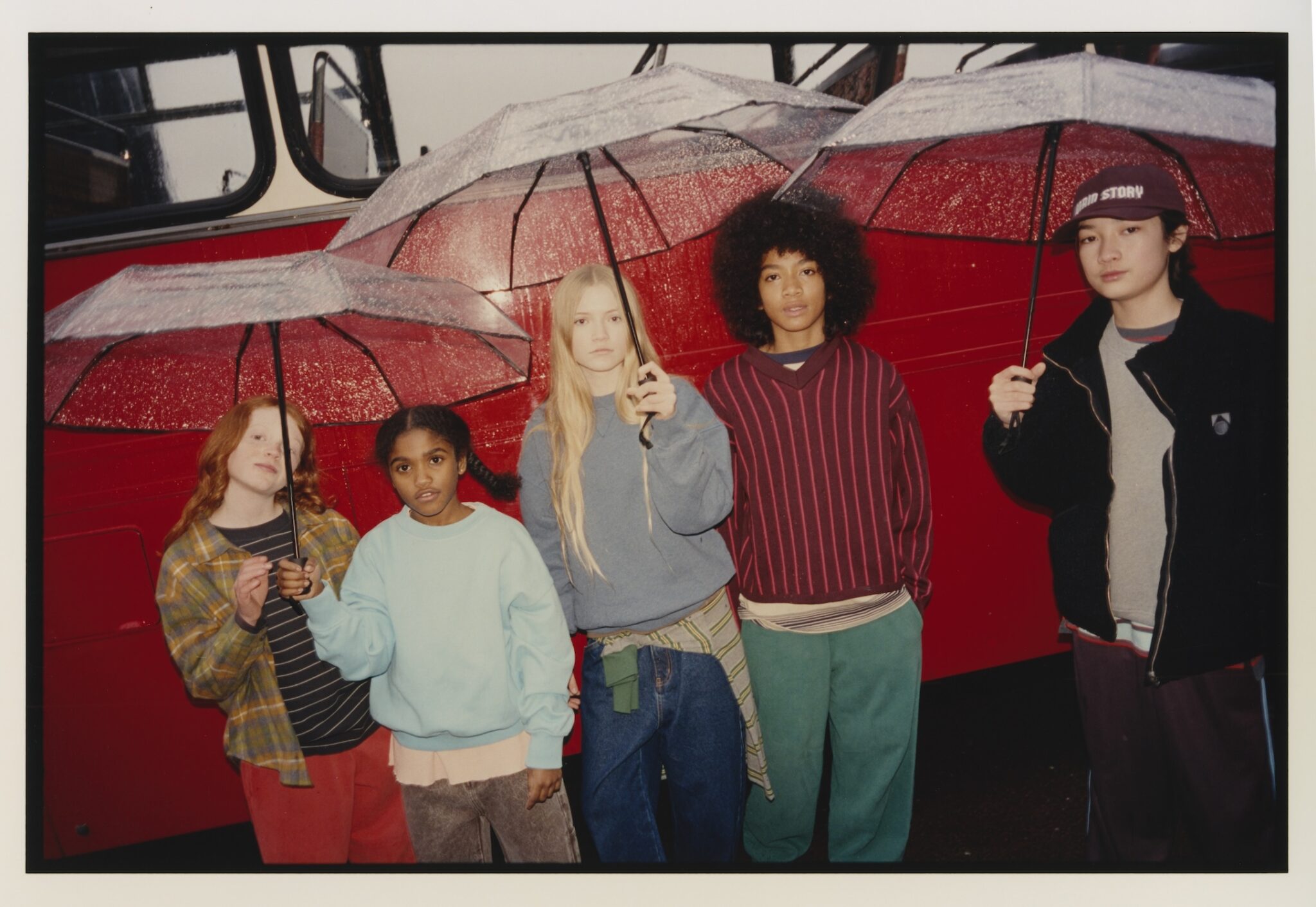 Five children wearing colourful knitwear and casual streetwear posing together by a canal in London.