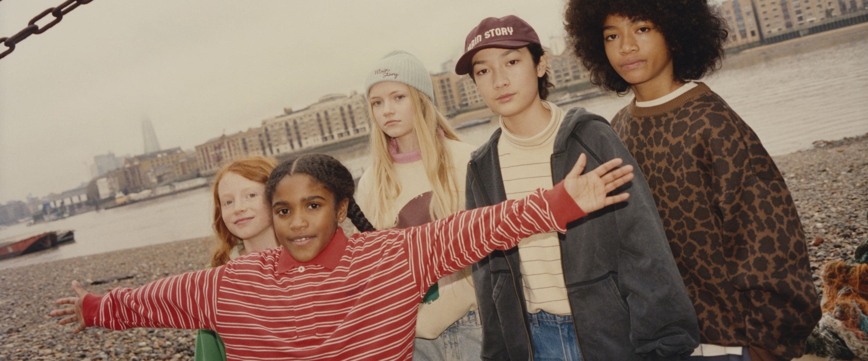Children in colourful casual outfits standing together by the River Thames, with London’s skyline in the background.