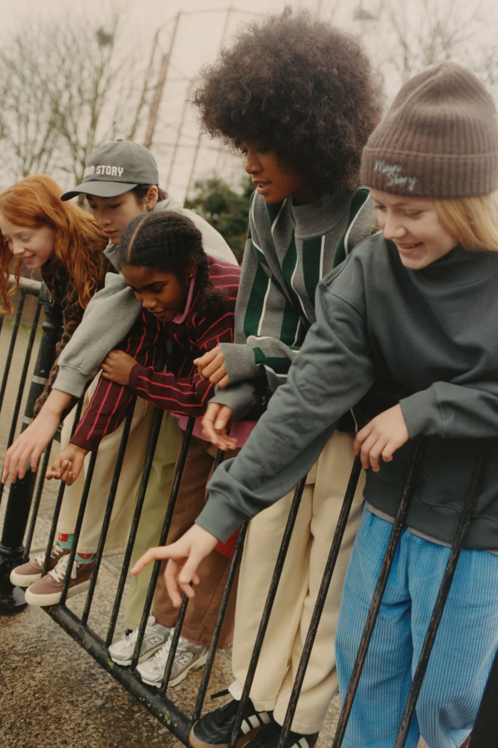 A group of children leaning over a riverside railing, wearing colourful Main Story knitwear and casual layers.
