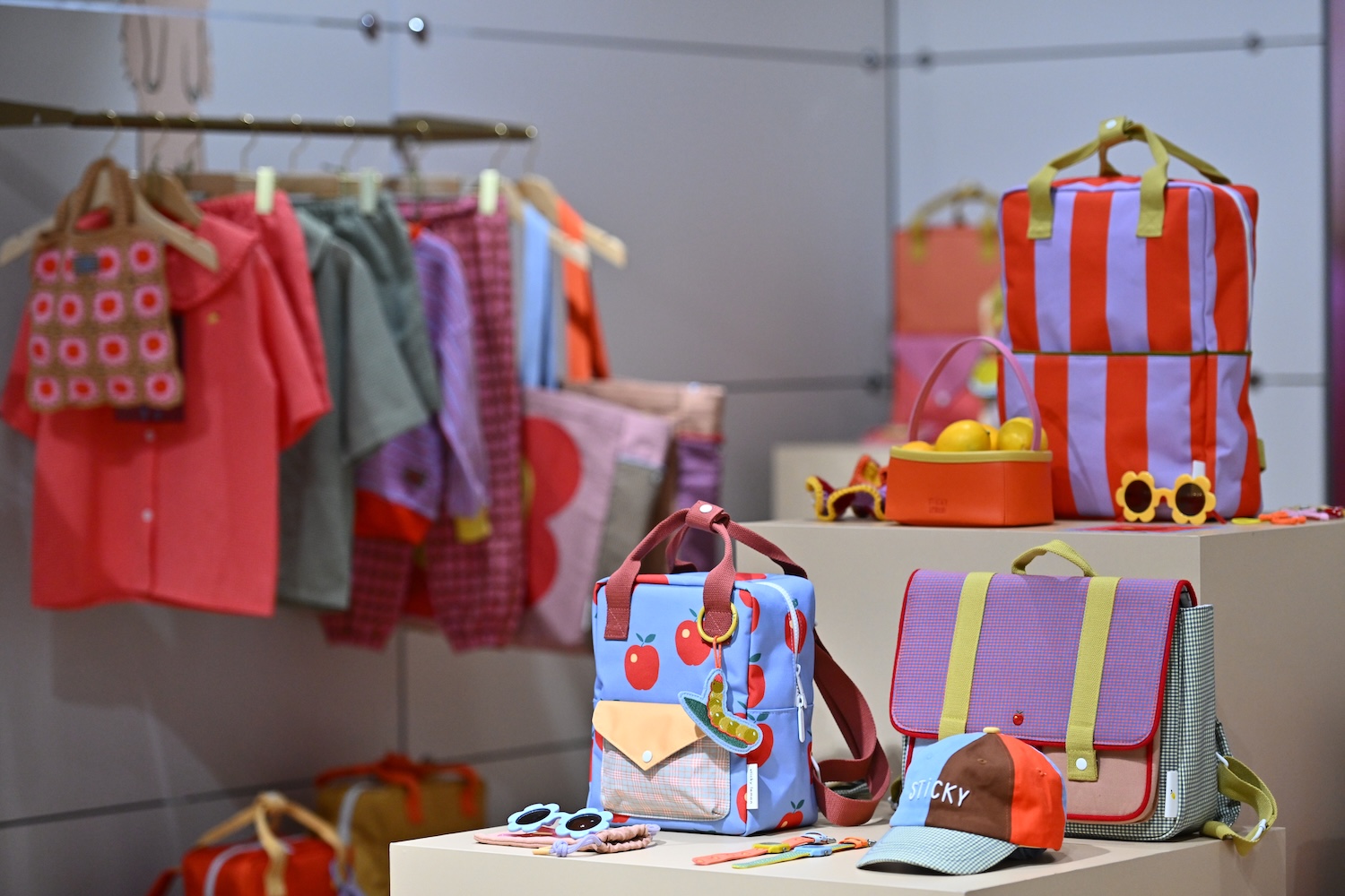 Colourful children’s fashion and accessories displayed at a kidswear trade fair, including backpacks, clothing, sunglasses and caps.