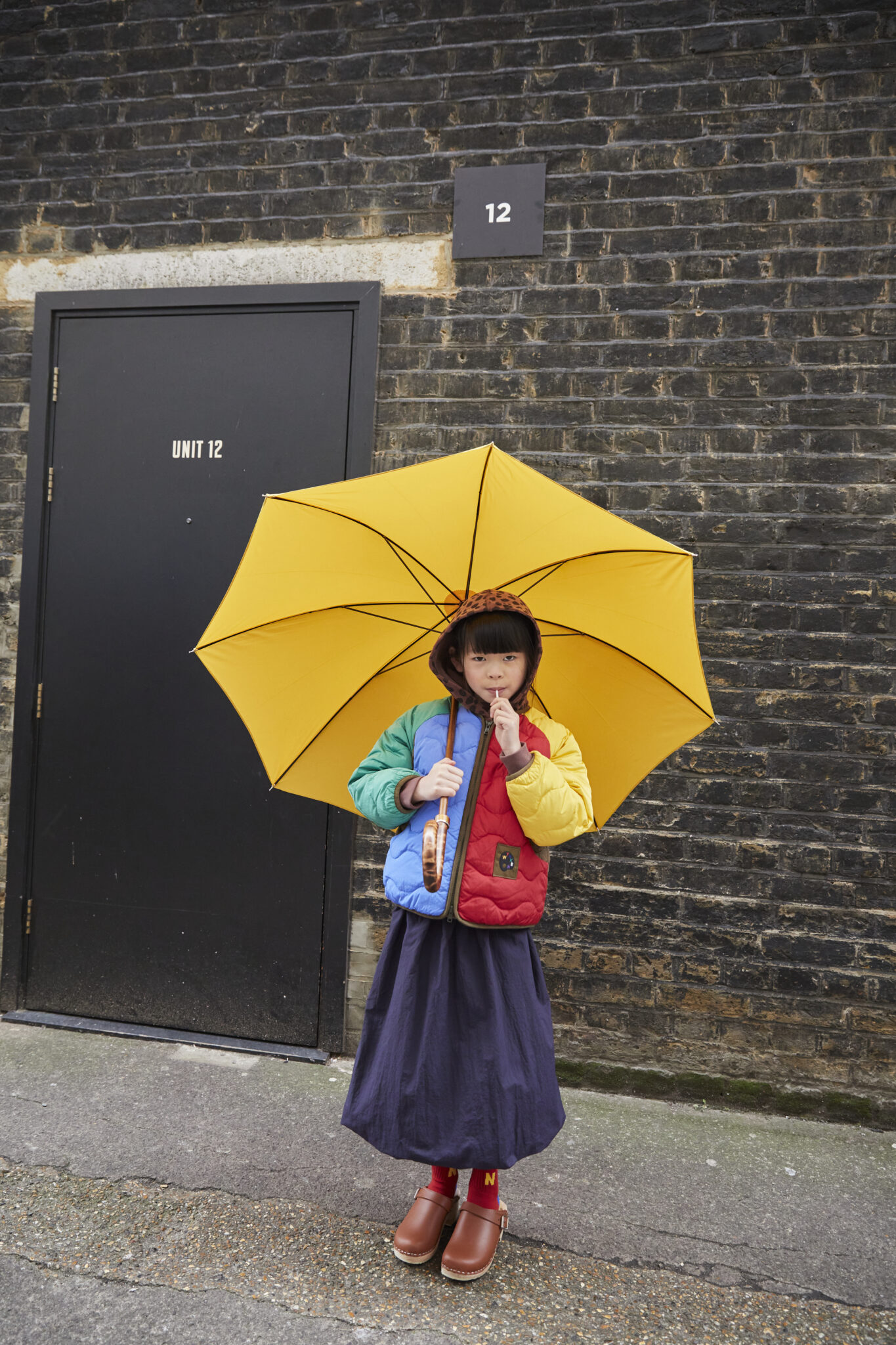 Wynken AW26 collection campaign featuring a child in a colourful quilted jacket, navy skirt and brown clogs holding a bright yellow umbrella.