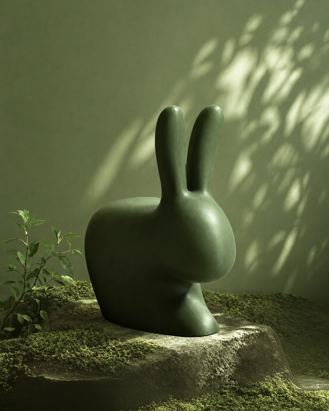Qeeboo’s Rabbit Chair in Deep Green, featuring its iconic rabbit-ear silhouette, set on a moss-covered stone pedestal against a sage-green backdrop with dappled sunlight.