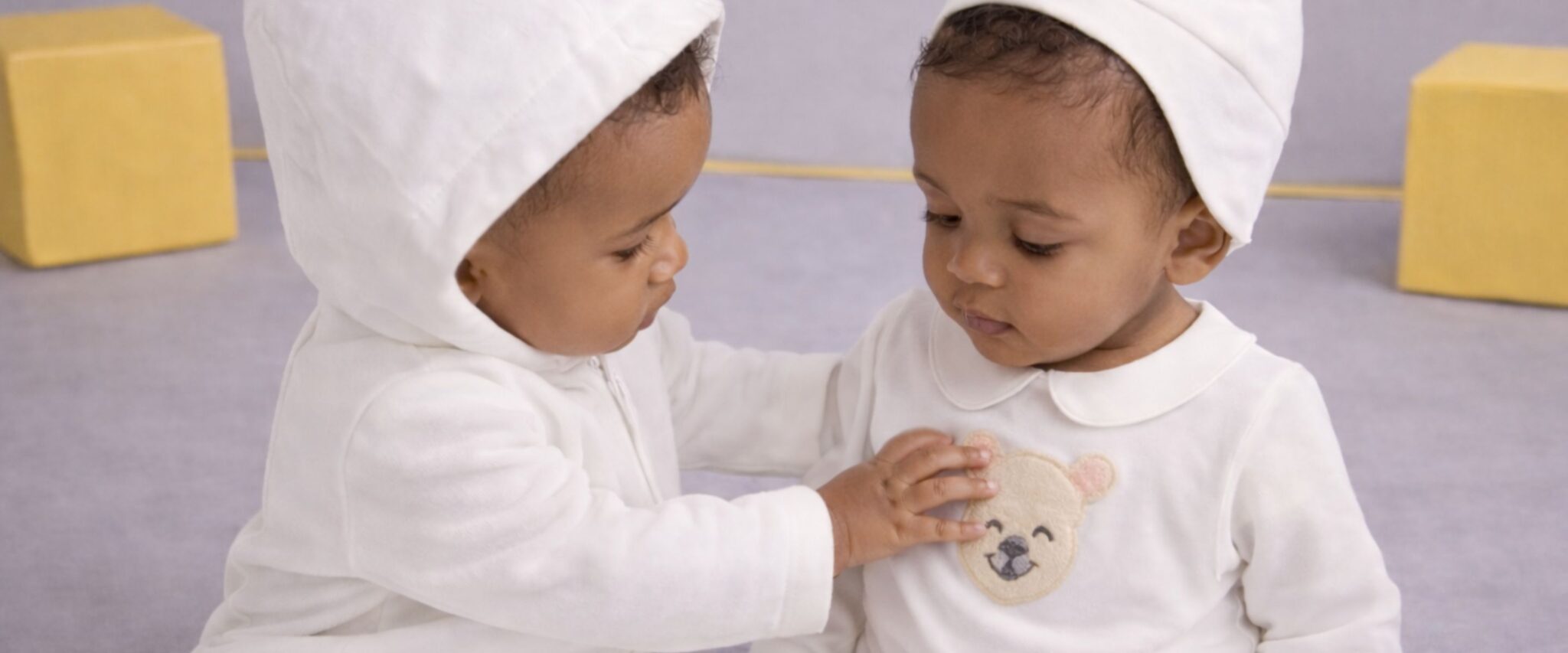 Two Gensami babies wearing white newborn outfits and bear-ear hats