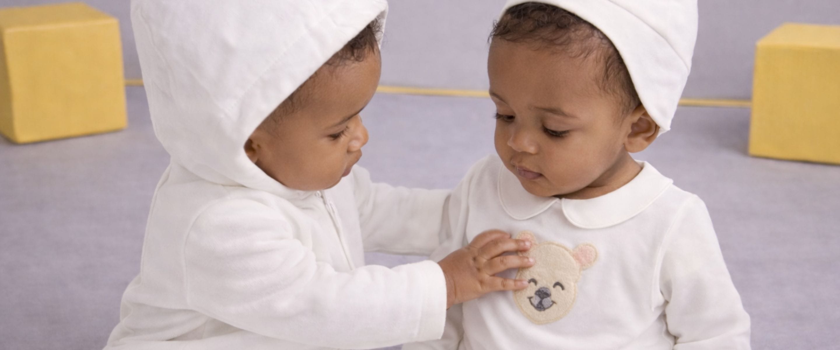 Two Gensami babies wearing white newborn outfits and bear-ear hats