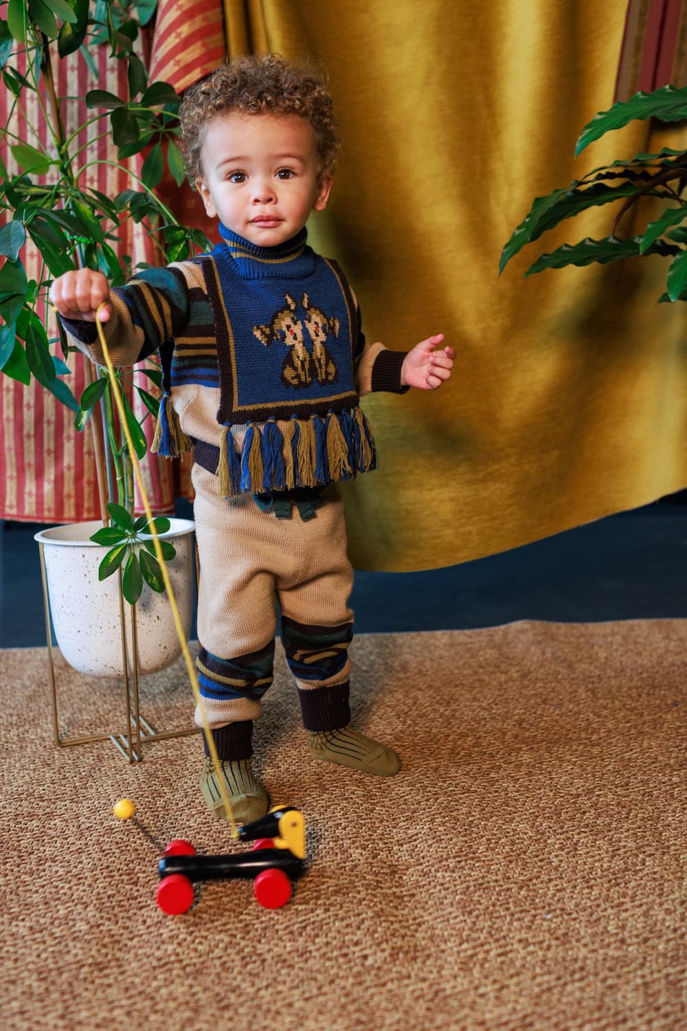 Toddler wearing a blue patterned knitted top with colourful fringe, beige trousers and striped socks from the Autumn/Winter 2026 collection.