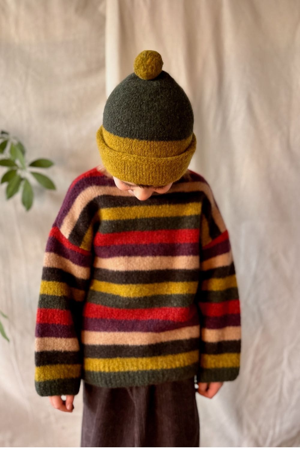 Child wearing a colourful striped knit sweater in red, mustard yellow, brown and green, paired with a matching mustard and grey beanie.