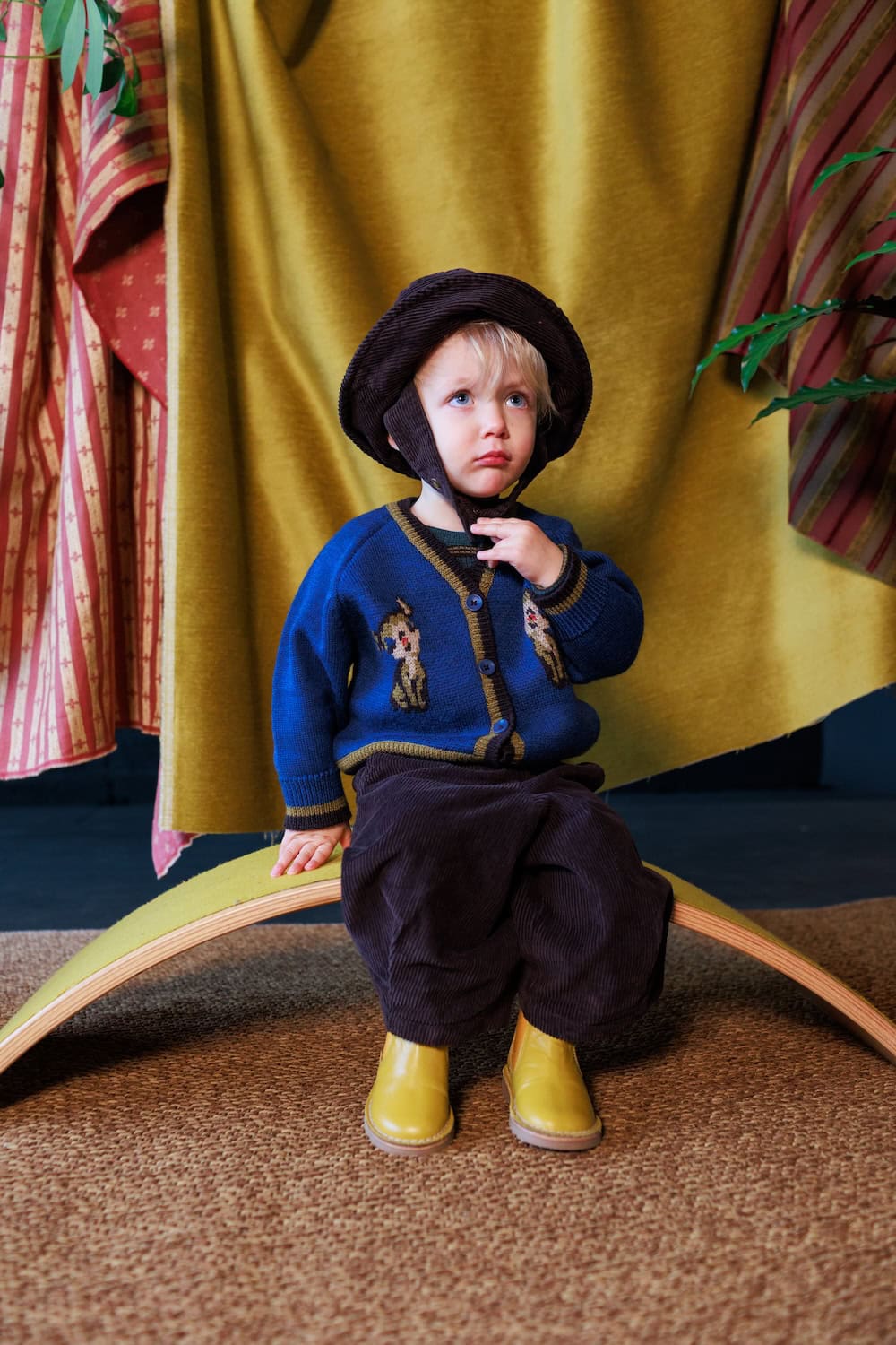 Toddler wearing a blue knitted cardigan with playful animal motifs, dark brown corduroy trousers and a vintage-inspired hat.