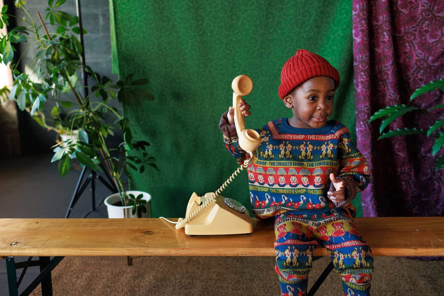 Child wearing a colourful patterned knitwear set featuring playful animal motifs and “The Curiosity Shop” lettering, styled with a red beanie and playing with a vintage telephone.