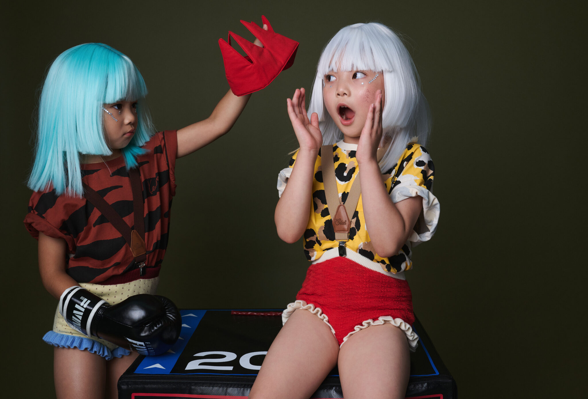 A girl in a turquoise wig lifts the red crown off her friend, who reacts with a surprised face