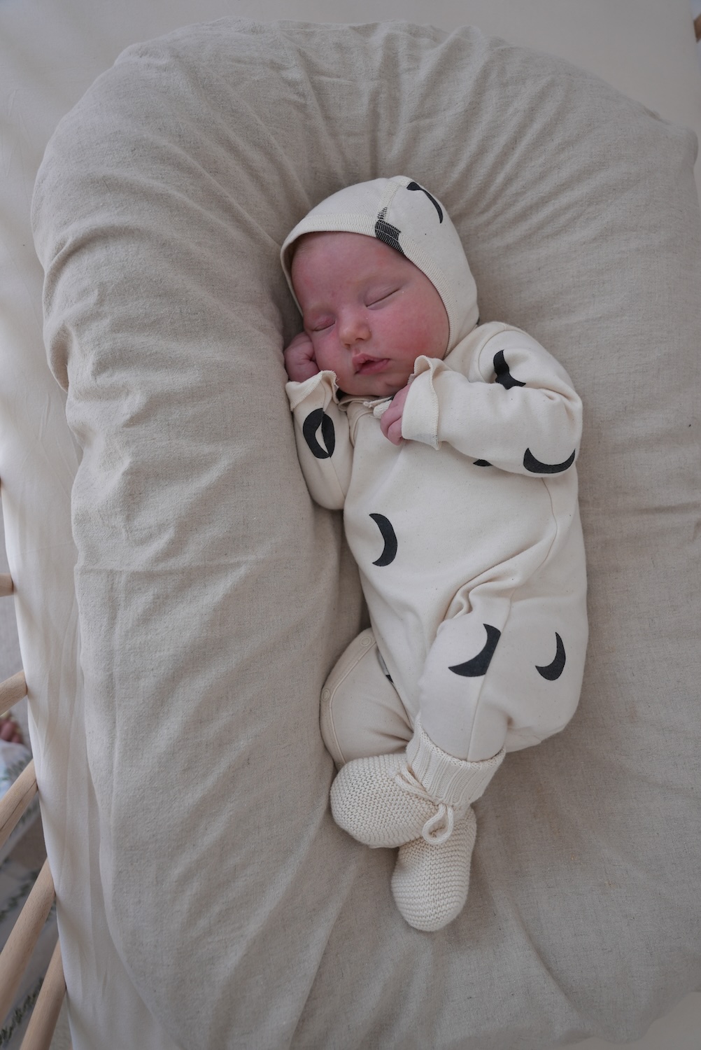 Sleeping newborn wearing a cream Organic Zoo x BINIBAMBA onesie and bonnet decorated with a black crescent moon print.