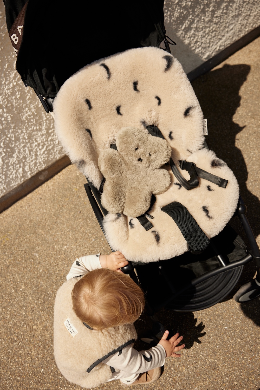 Child sitting in a stroller with a moon-print sheepskin liner, while another child wearing a matching vest reaches towards it.