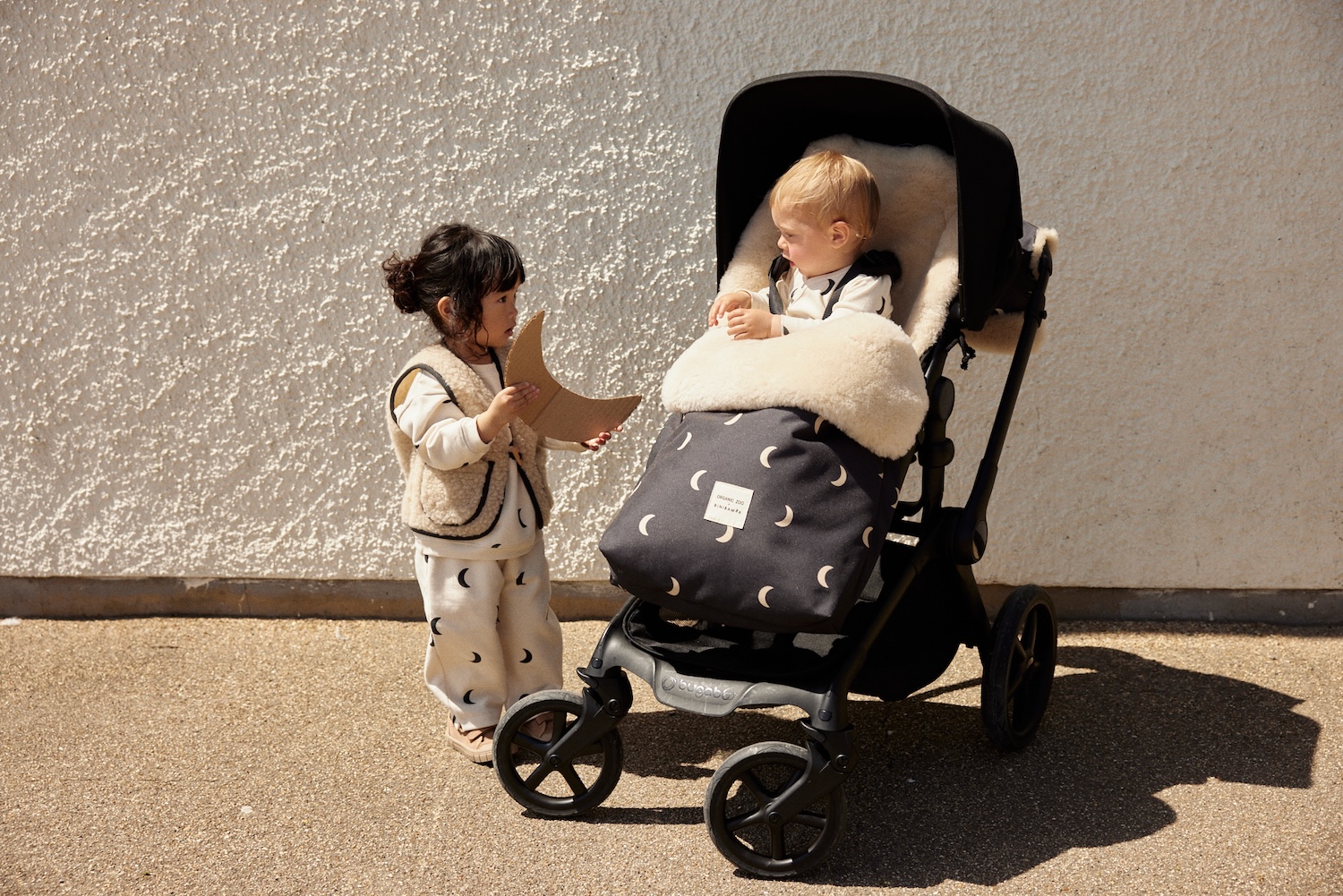 Two children wearing Organic Zoo x BINIBAMBA Midnight outfits beside a stroller with a moon-print sheepskin liner and footmuff.