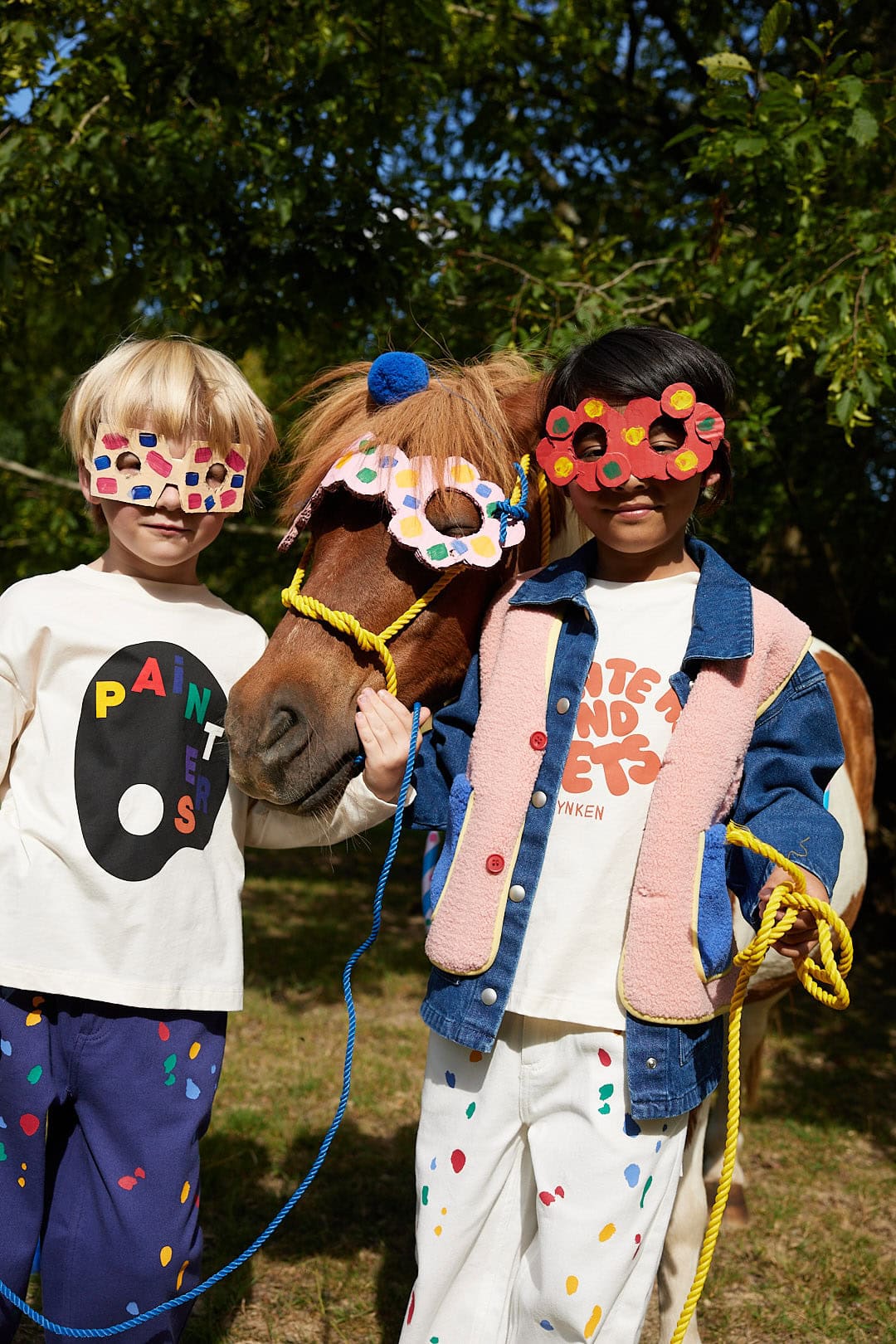 Children wearing Wynken AW26 Painters and Poets collection, posing with a horse wearing a colourful handmade mask. Playful painted masks, graphic T-shirts and colourful trousers celebrate creativity and childhood imagination.