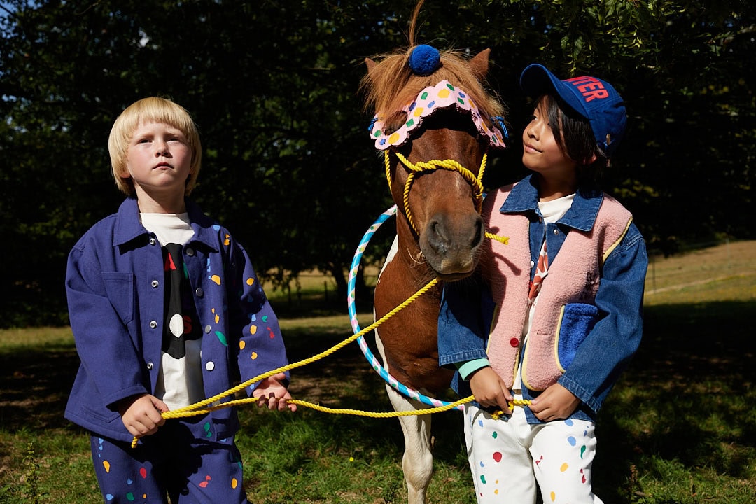 Wynken AW26 Painters and Poets collection featuring two children with a horse outdoors. Colourful graphic T-shirts, blue jackets, a pink fleece-trimmed denim vest and playful accessories bring the collection’s artistic spirit to life.