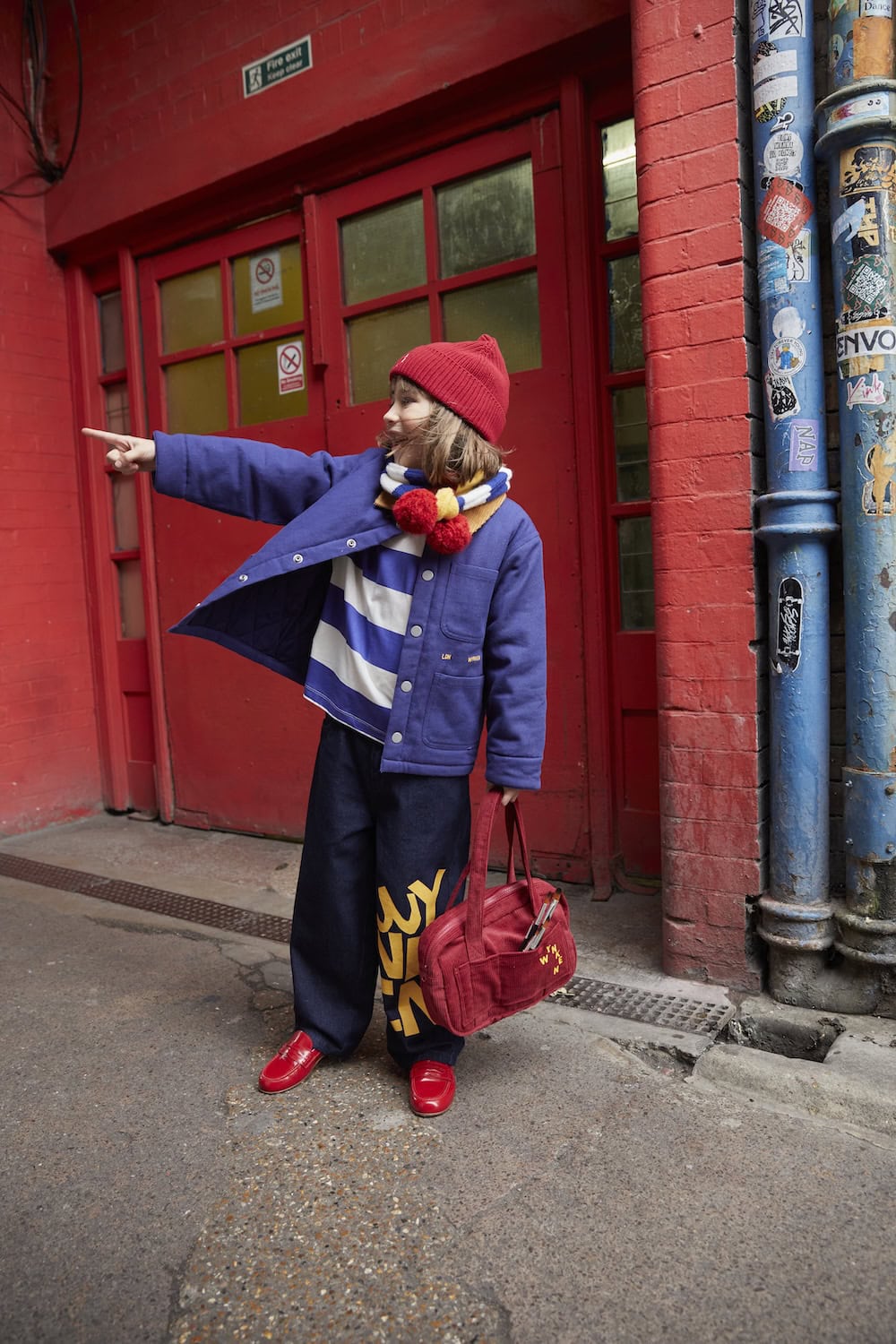 Wynken AW26 kidswear collection featuring a child in a cobalt blue jacket, striped top, red beanie, colourful pom-pom scarf and wide-leg jeans with bold yellow lettering, carrying a red bag against a vibrant red urban backdrop.