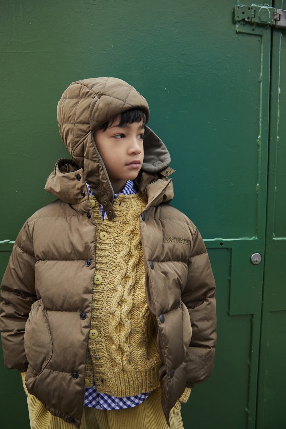 Wynken AW26 kidswear collection featuring a child wearing a taupe quilted puffer jacket with a matching earflap hood, a mustard cable-knit sweater and a blue gingham shirt, styled against a green door.