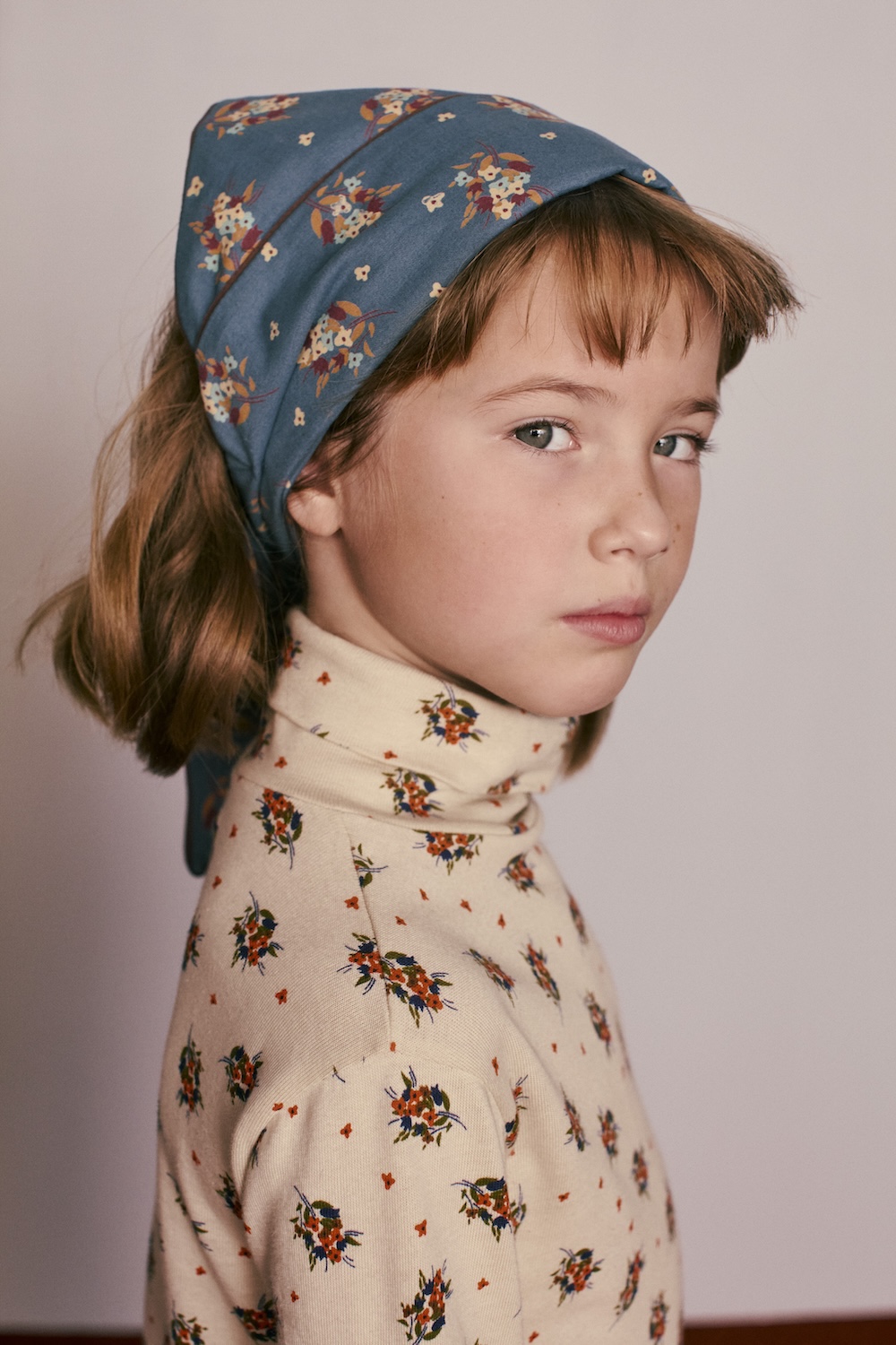 Close-up of a child wearing a blue floral headscarf and a cream turtleneck with delicate floral prints from Caramel for Zara.